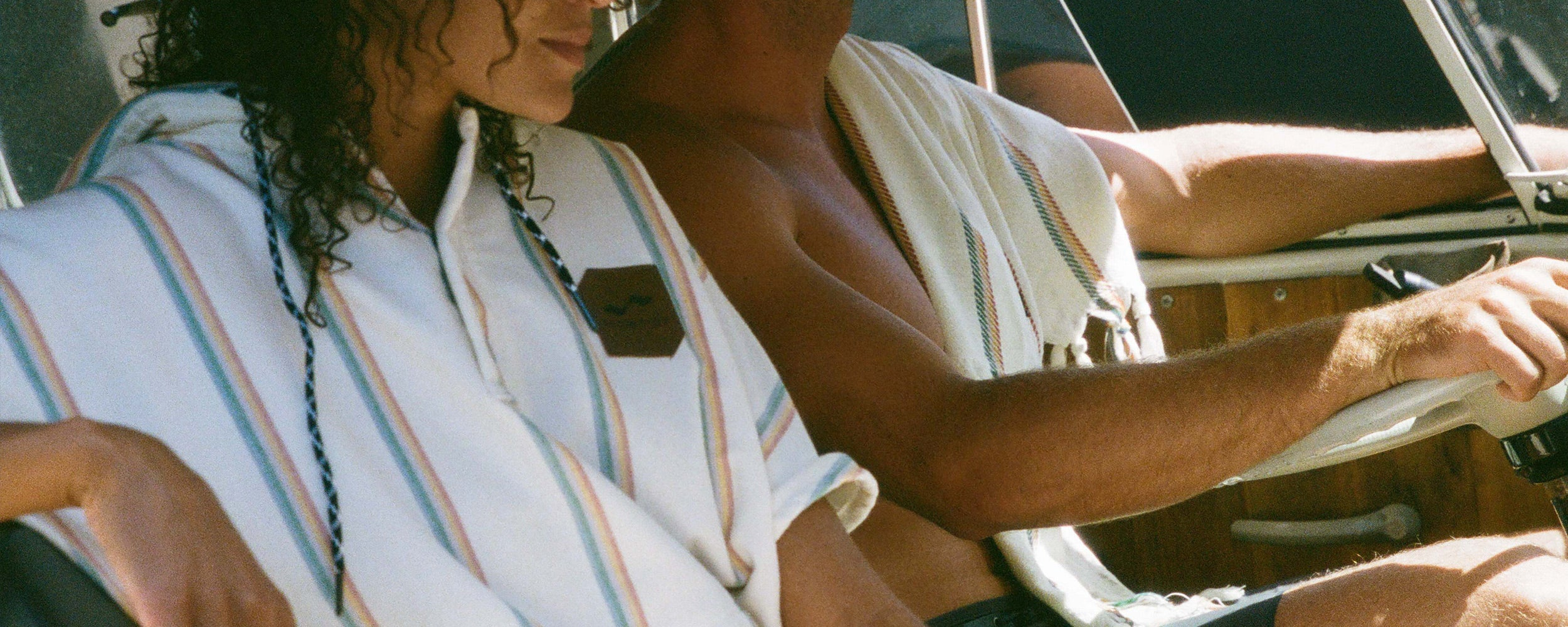 Two people sit side by side in a boat, one wearing a striped poncho towel and the other shirtless with a towel draped over his shoulder. Both appear relaxed and are enjoying the moment.