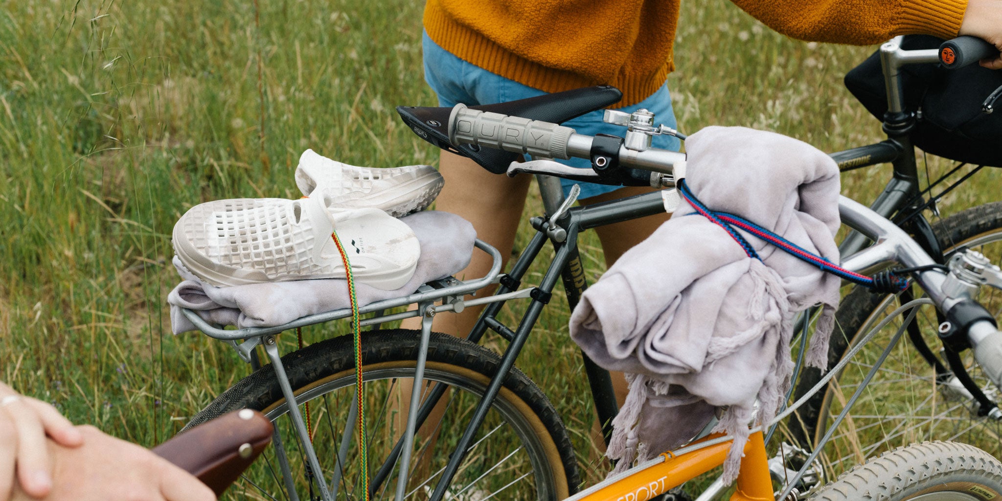 A person stands by an orange bike with a gray blanket and white sneakers strapped to the back rack, on a grassy field. Only their legs and hand are visible. Another bike is partially seen.