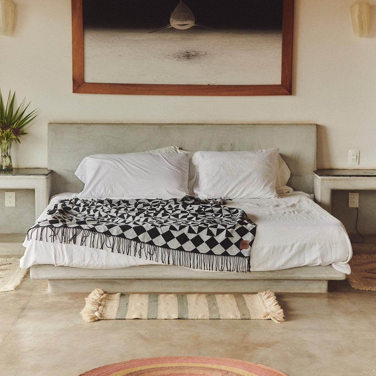 A modern bed with white linens and a black-and-white geometric throw sits against a concrete headboard. A shark photo hangs above, with small plants and shelves on either side of the bed.
