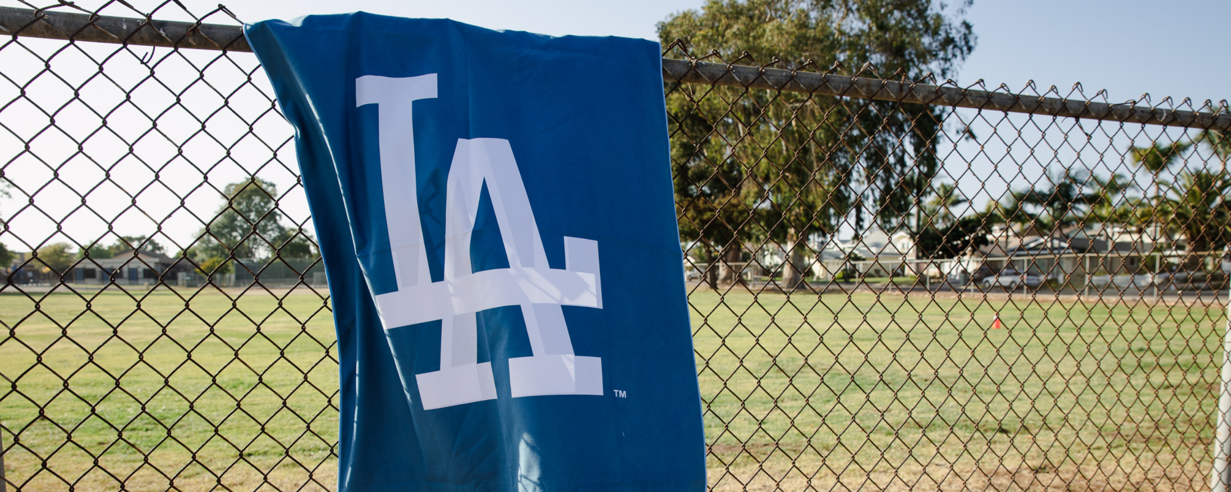A blue flag with a large white LA logo hangs on a chain-link fence, with a grassy field and trees visible in the background.