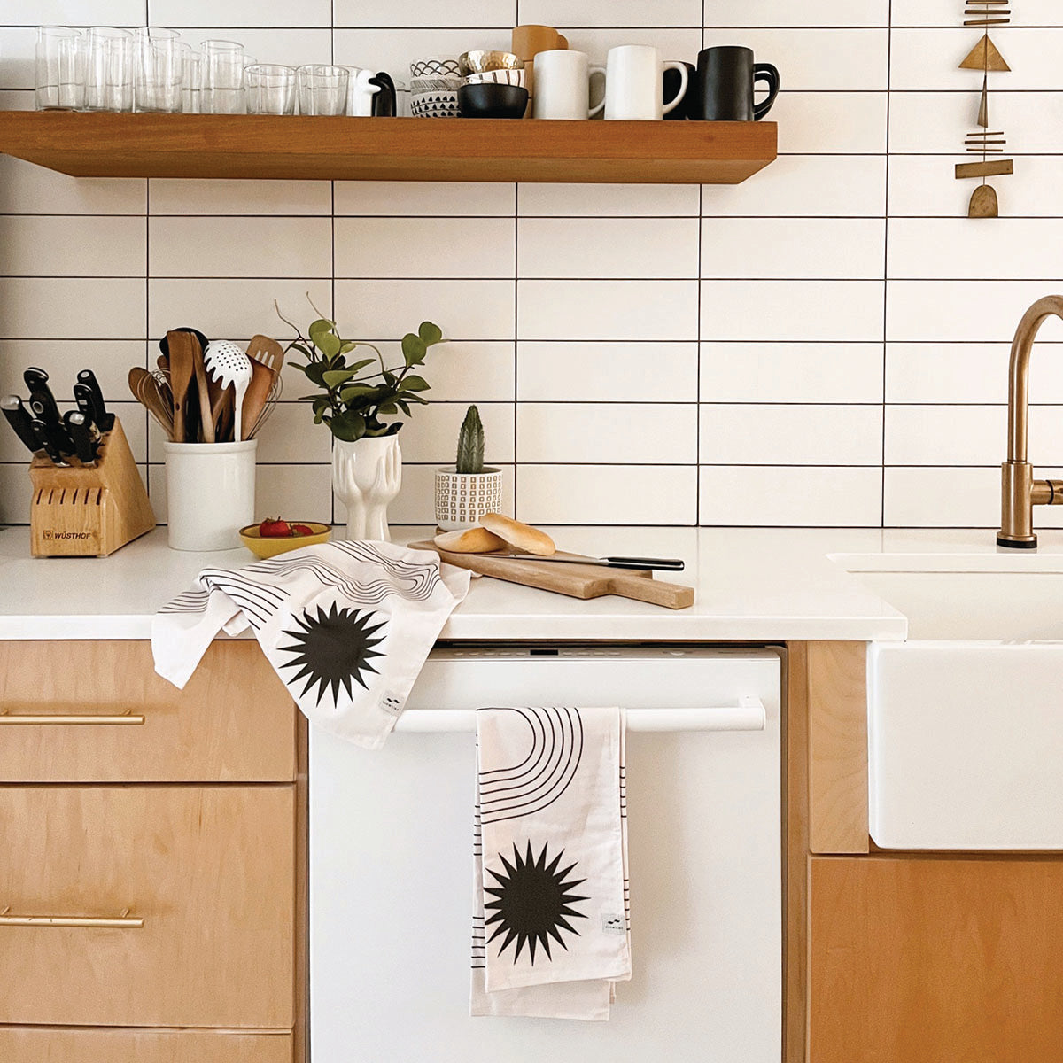 Modern kitchen with light wood cabinets, white tile backsplash, open shelf with dishes, utensils in containers, a plant, and dish towels with black sunburst patterns draped on the counter and dishwasher.