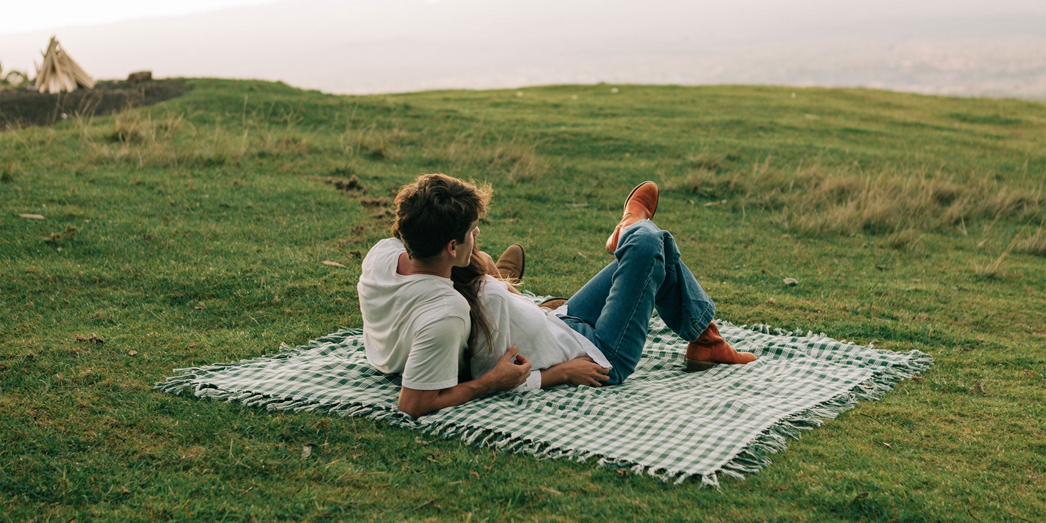 A couple lies on a checkered blanket on grass, relaxing and looking at the landscape. They are casually dressed, and the scene is peaceful with nature in the background.