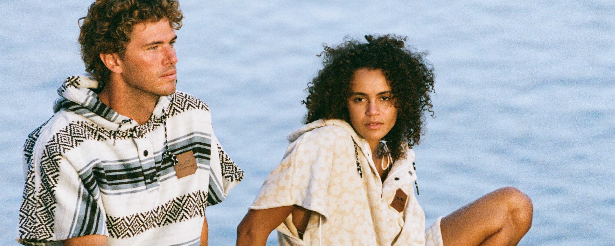 A man and a woman wearing patterned hooded ponchos sit by the water, gazing thoughtfully into the distance.