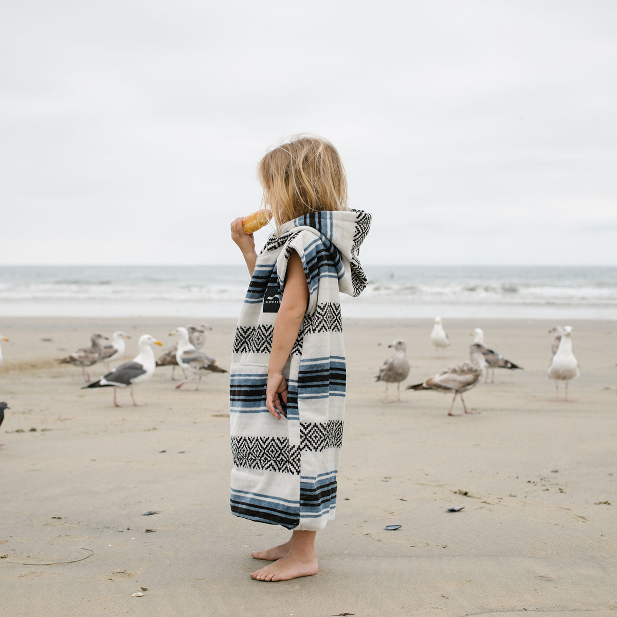 A young child in a hooded towel stands barefoot on a sandy beach, eating an ice cream cone. Seagulls gather in the background with the ocean and cloudy sky behind them.