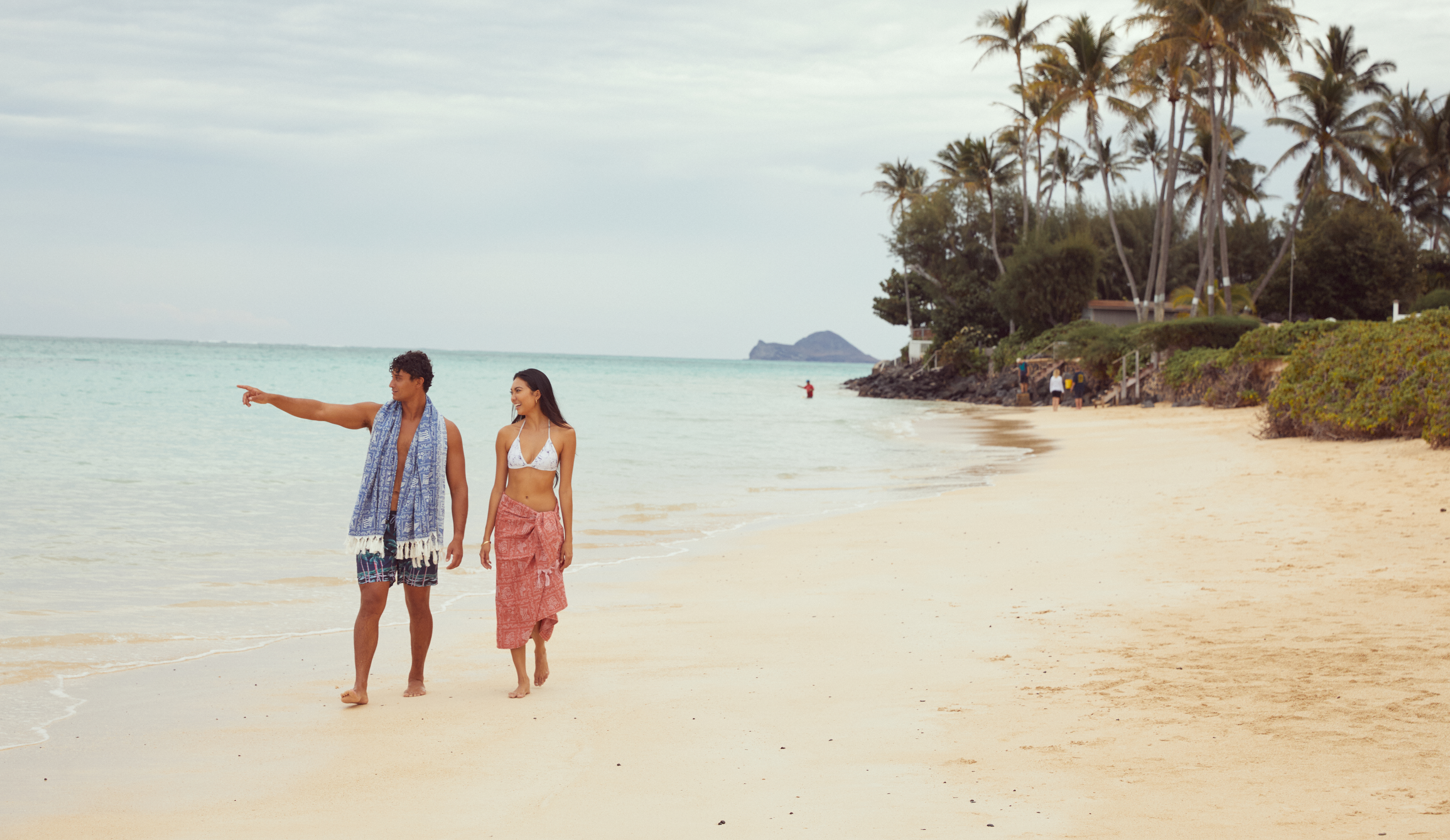 A man and woman walk along a sandy beach. The man points toward the ocean while talking. Palm trees and greenery line the shore, and a person is seen in the distance near the water.