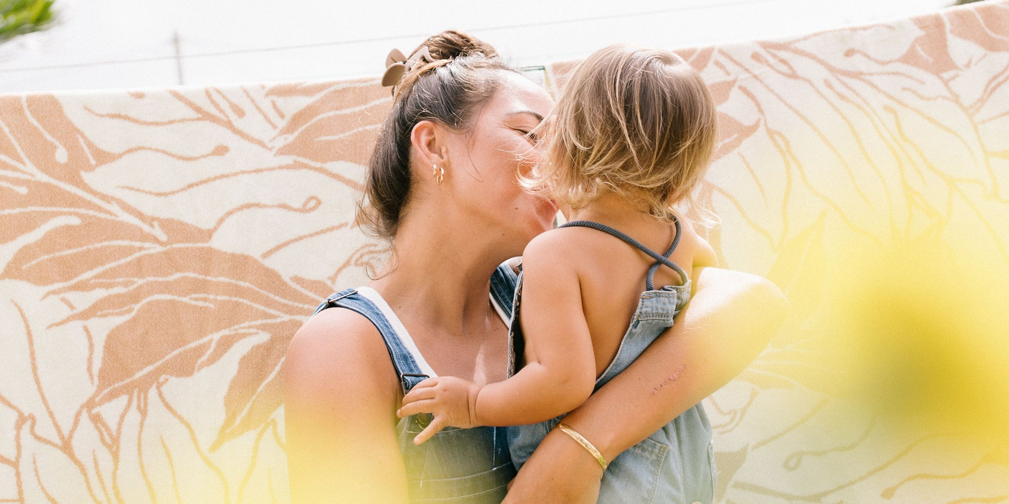 A woman in a denim outfit holds and kisses a small child, who is also wearing denim. They stand outdoors in front of a patterned cloth, with soft yellow flowers in the foreground.