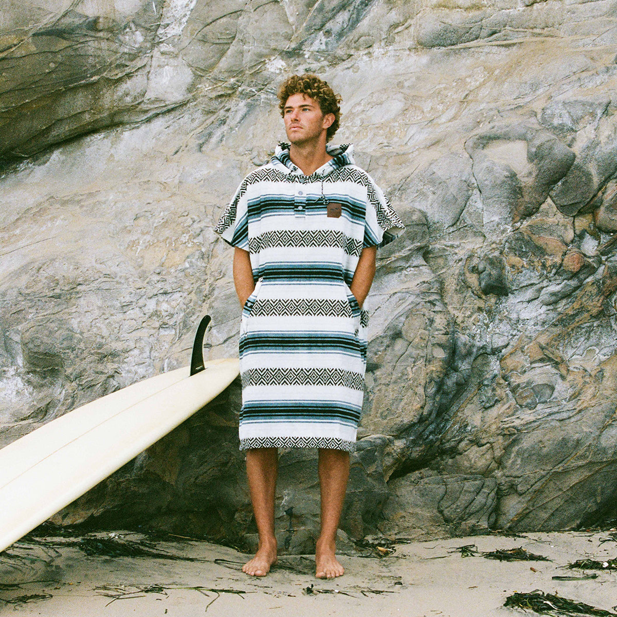 A man in a striped poncho stands barefoot on the sand near a rock wall, with a surfboard propped up beside him.