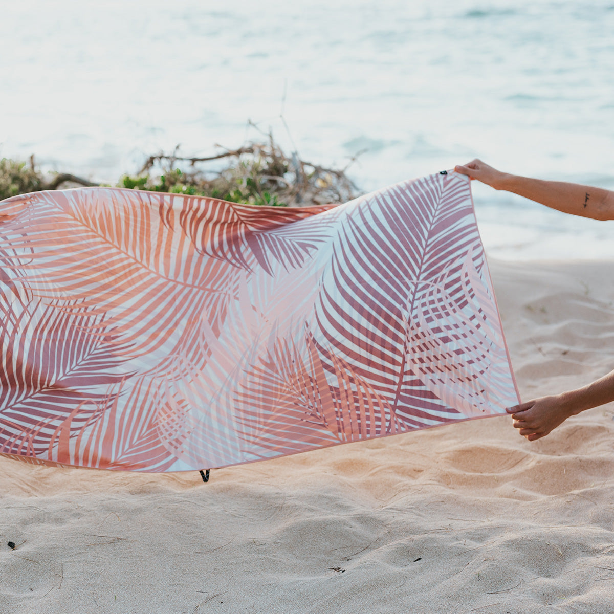 Person holding a pink and orange palm leaf-patterned towel on a sandy beach near the ocean, with waves and driftwood visible in the background.