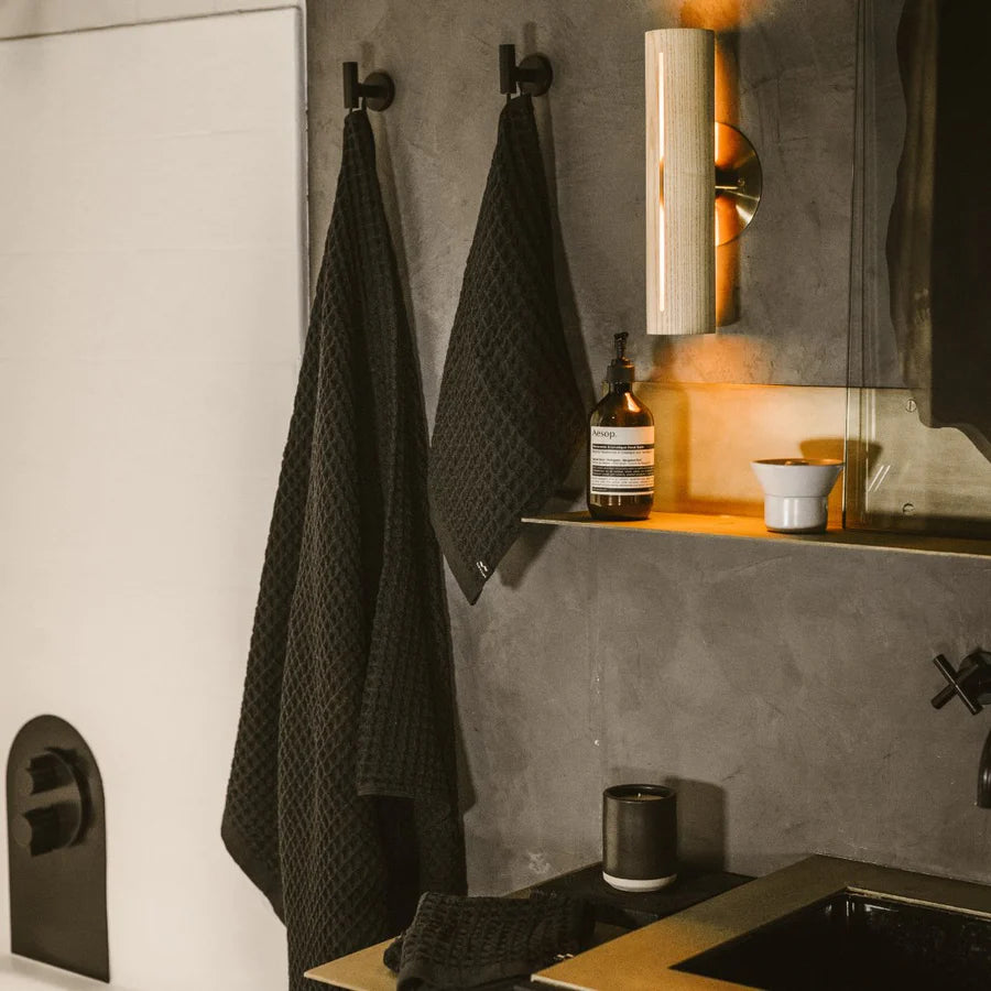 A modern bathroom with two black textured towels hanging on hooks, a gold wall sconce emitting warm light, a bottle of soap, a small cup, and a black sink with a folded towel nearby.