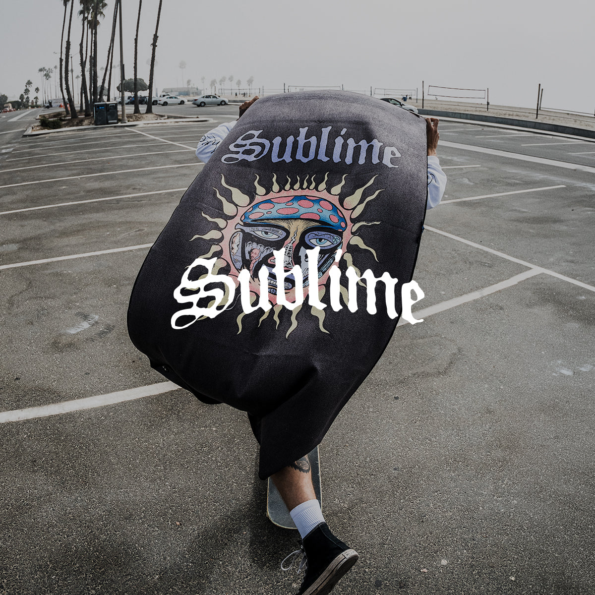 A person walking in a parking lot by the beach holds a large blanket with a colorful sun face and the word Sublime on it. The word Sublime is also overlaid in white text.