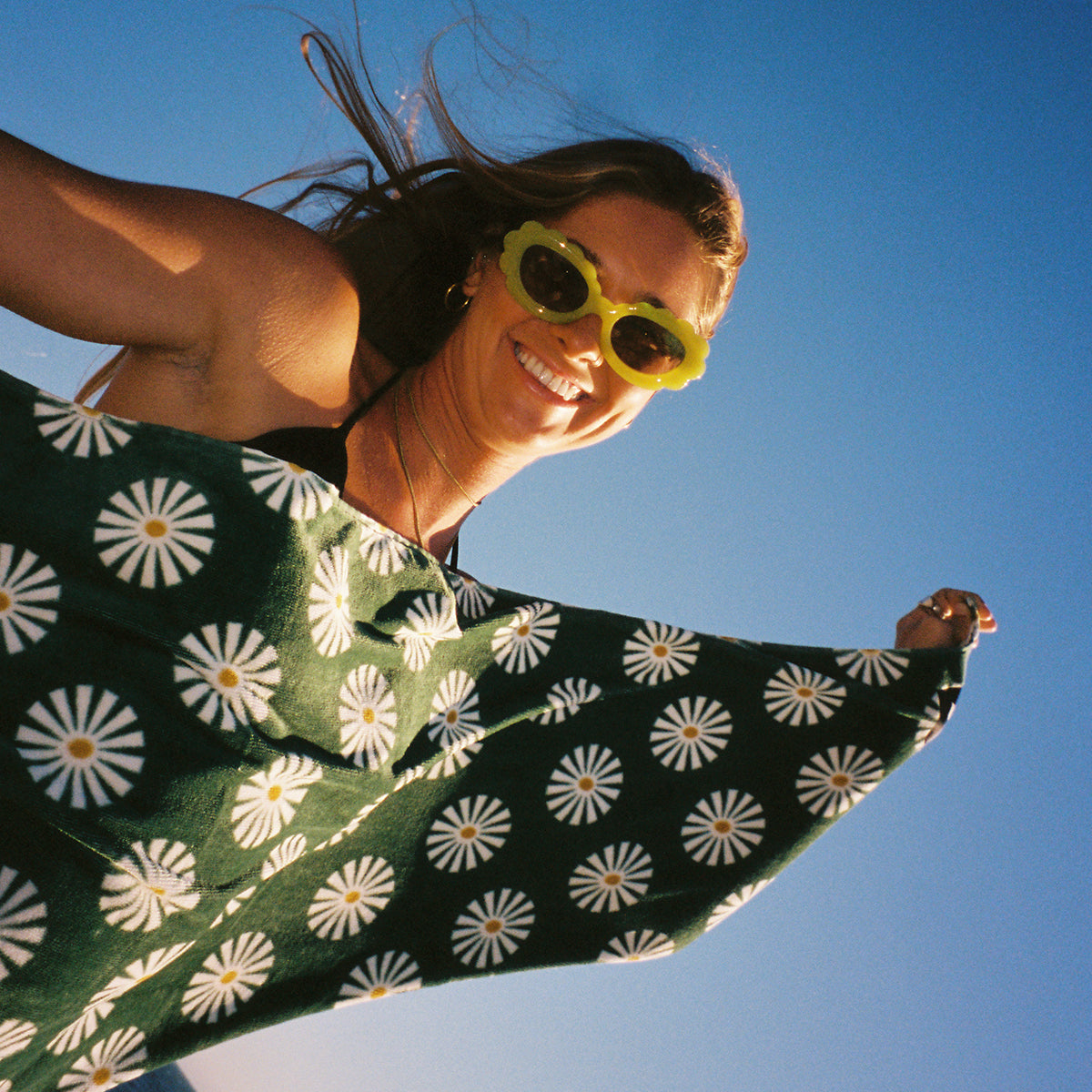 A smiling woman wearing yellow flower-shaped sunglasses holds out a green shawl with white daisy patterns against a clear blue sky.