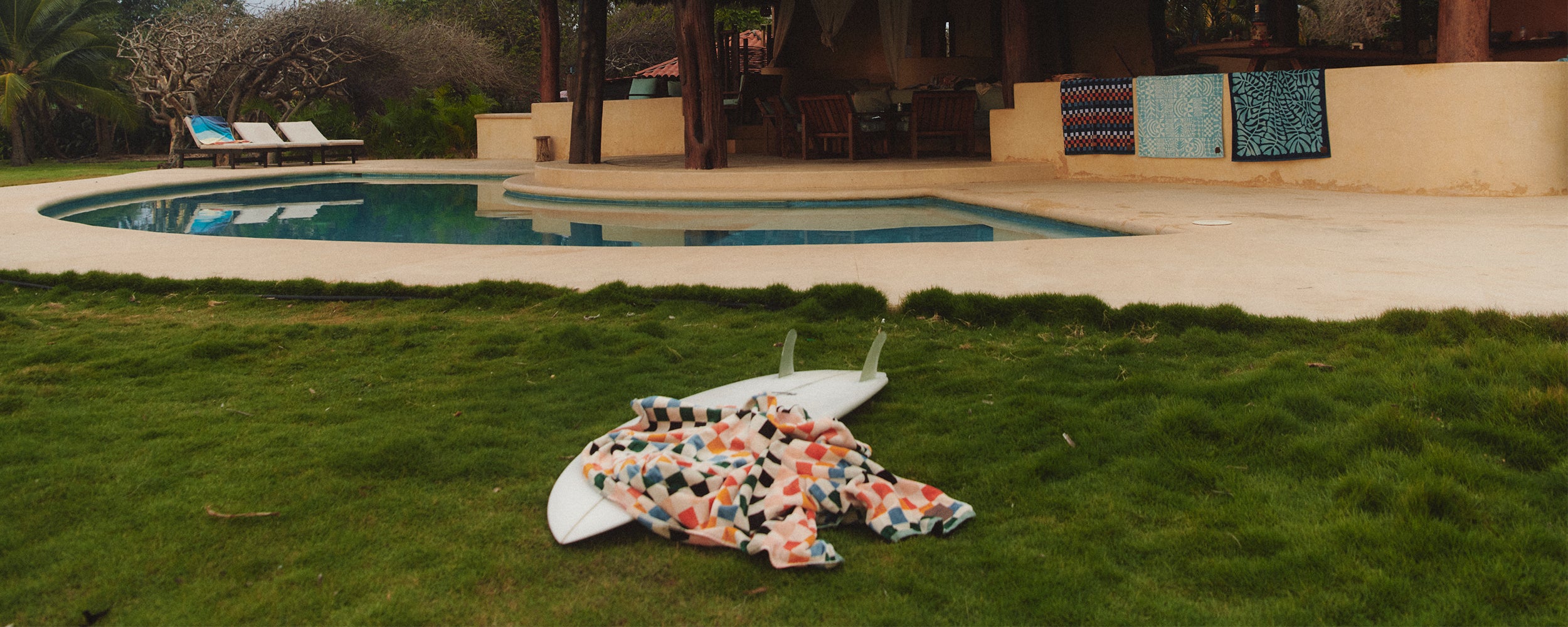 A surfboard and colorful towel lie on grass near a curvy pool, with lounge chairs and patterned towels hanging by a covered patio in the background.