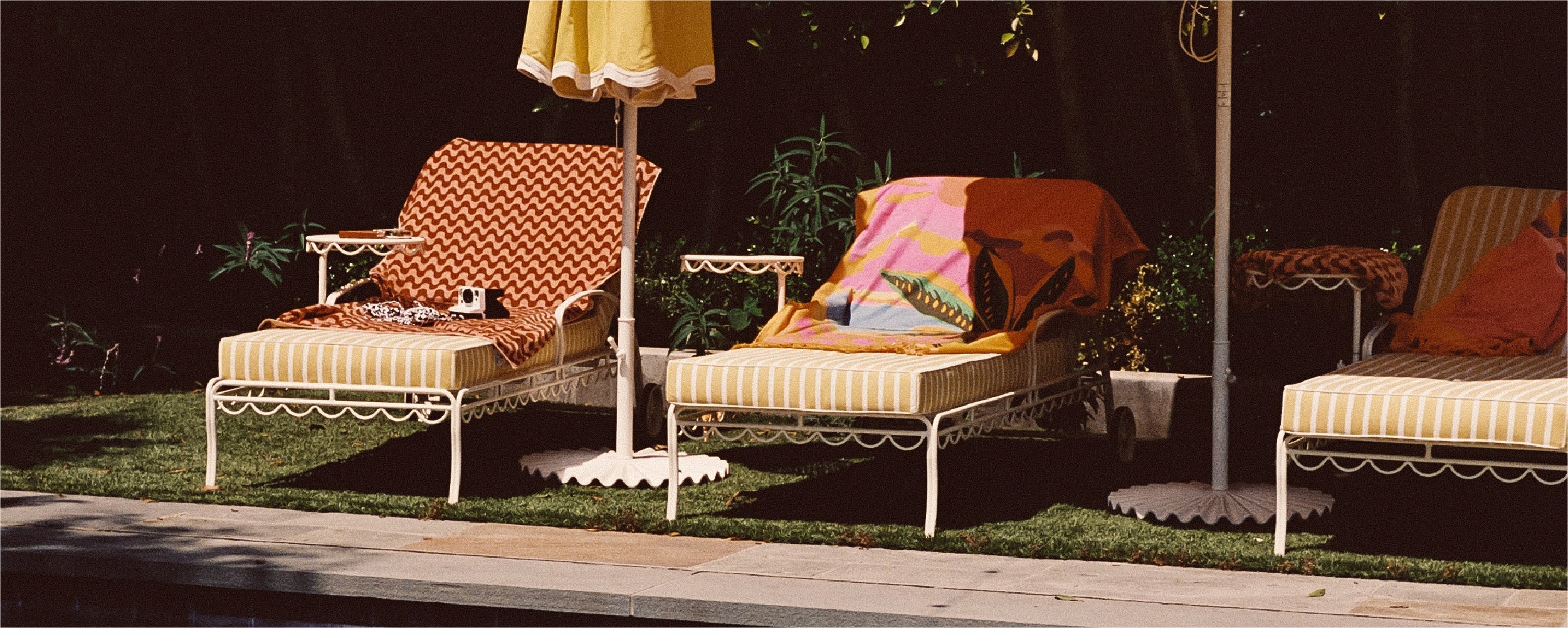 Two striped poolside lounge chairs with colorful towels and a cat resting on one chair, shaded by umbrellas, sit on grass next to a stone poolside, surrounded by greenery.
