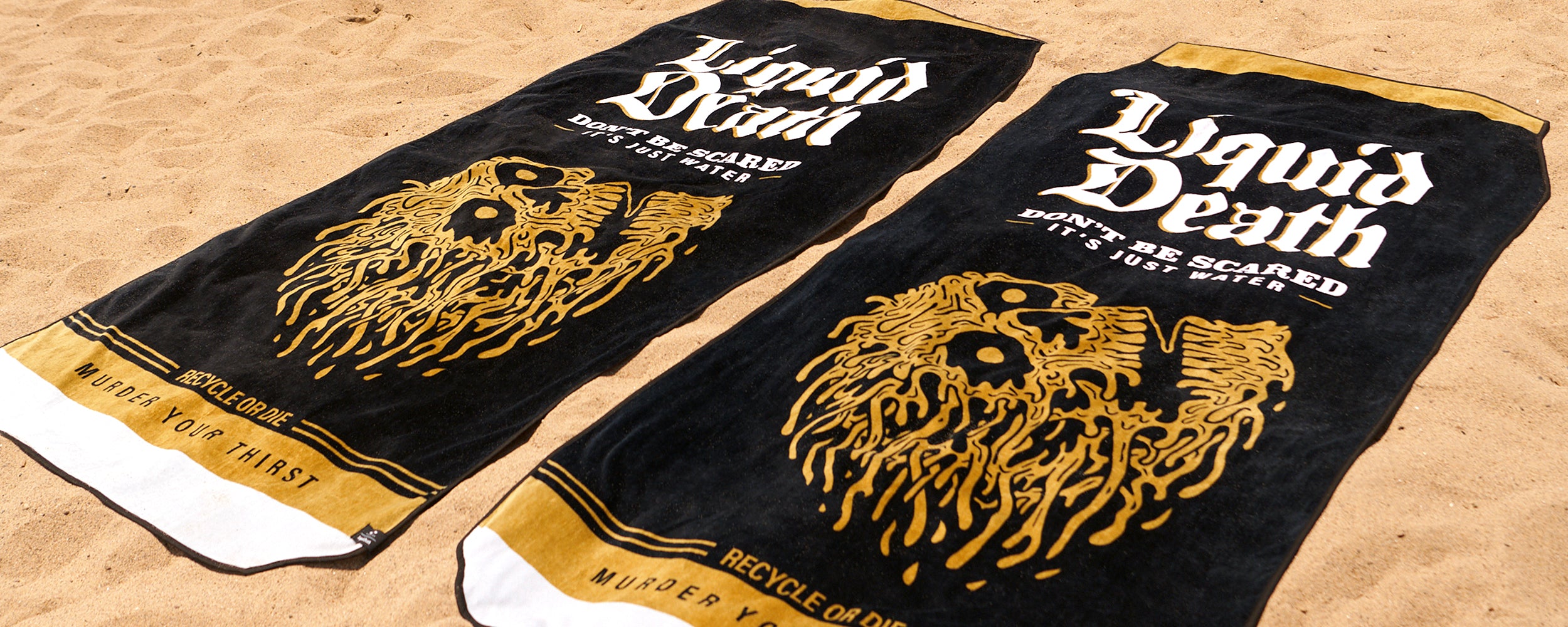 Two black and gold beach towels with Liquid Death, Dont Be Scared, Its Just Water and a gold skull illustration, laid out on sandy beach. Text at bottom reads Recycle or Die and Murder Your Thirst.