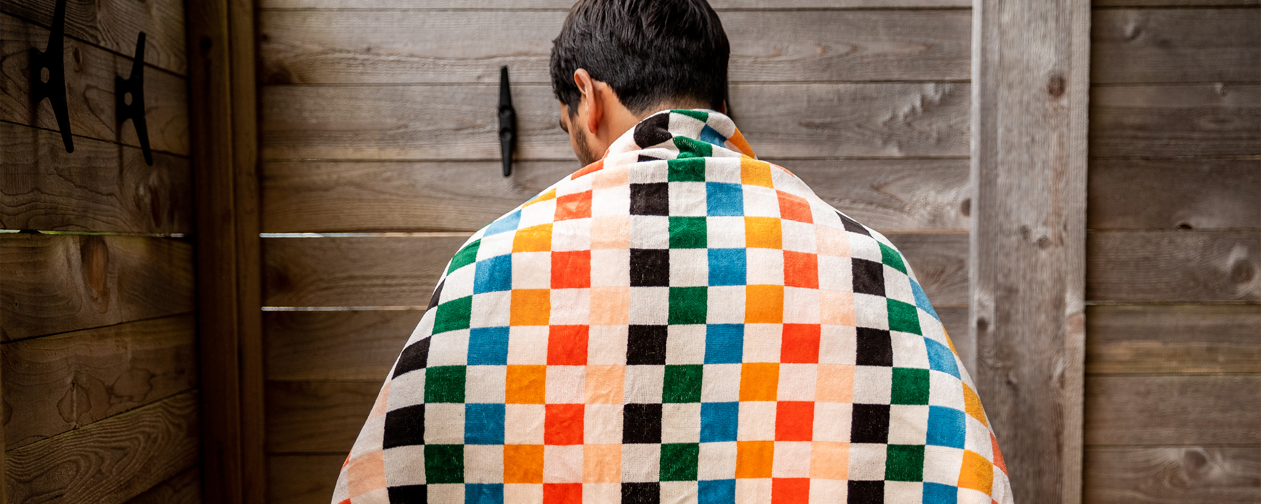 A person with short dark hair is seen from behind, wrapped in a colorful checkered towel, standing next to a wooden fence.