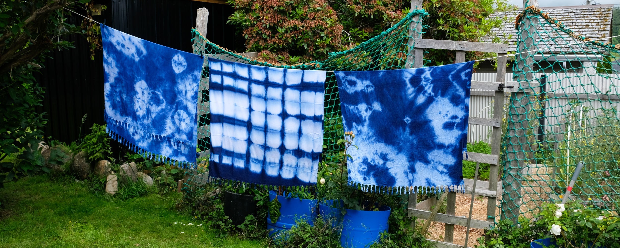 Three blue and white tie-dye fabrics hang on a green net clothesline outdoors, with plants, a wooden fence, and blue buckets in the background.