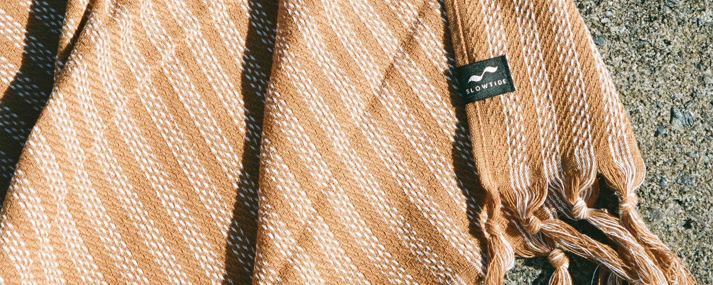 A close-up of a tan and white woven scarf with fringe ends, laying on a textured surface. A small black tag with SLOWTIDE and a wave logo is attached to the scarf.