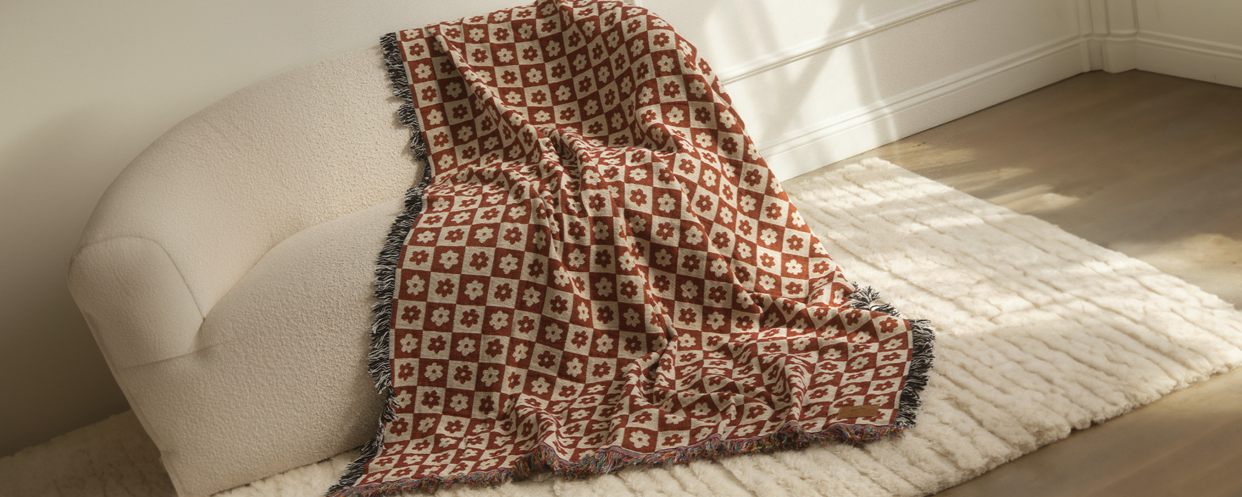 A cream-colored armchair draped with a red and cream checkered throw blanket featuring floral patterns, set on a fluffy cream rug in a sunlit room with hardwood floors.