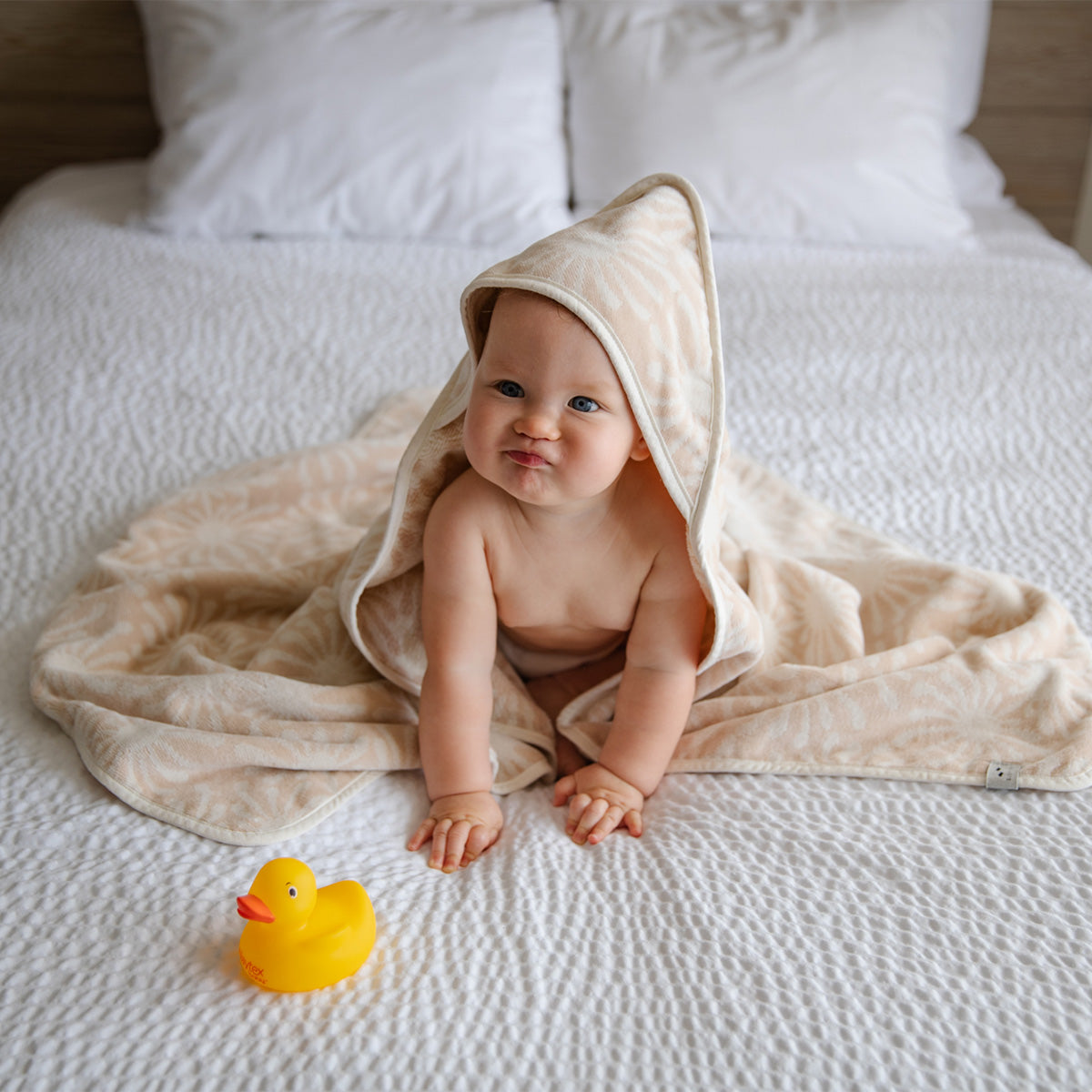 A baby wrapped in a beige hooded towel sits on a white bed, making a funny face. A yellow rubber duck is placed in front of the baby.