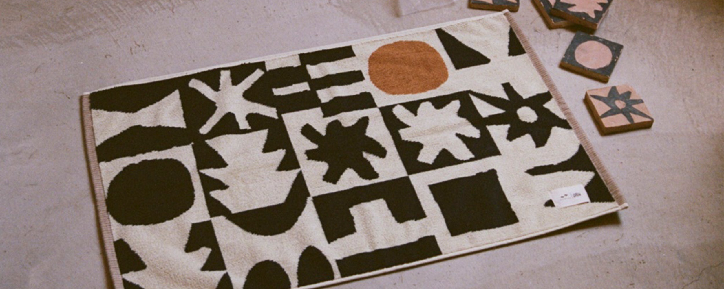A rectangular rug with abstract black shapes and one orange circle on a cream background lies on a concrete floor. Four patterned tiles are scattered nearby.