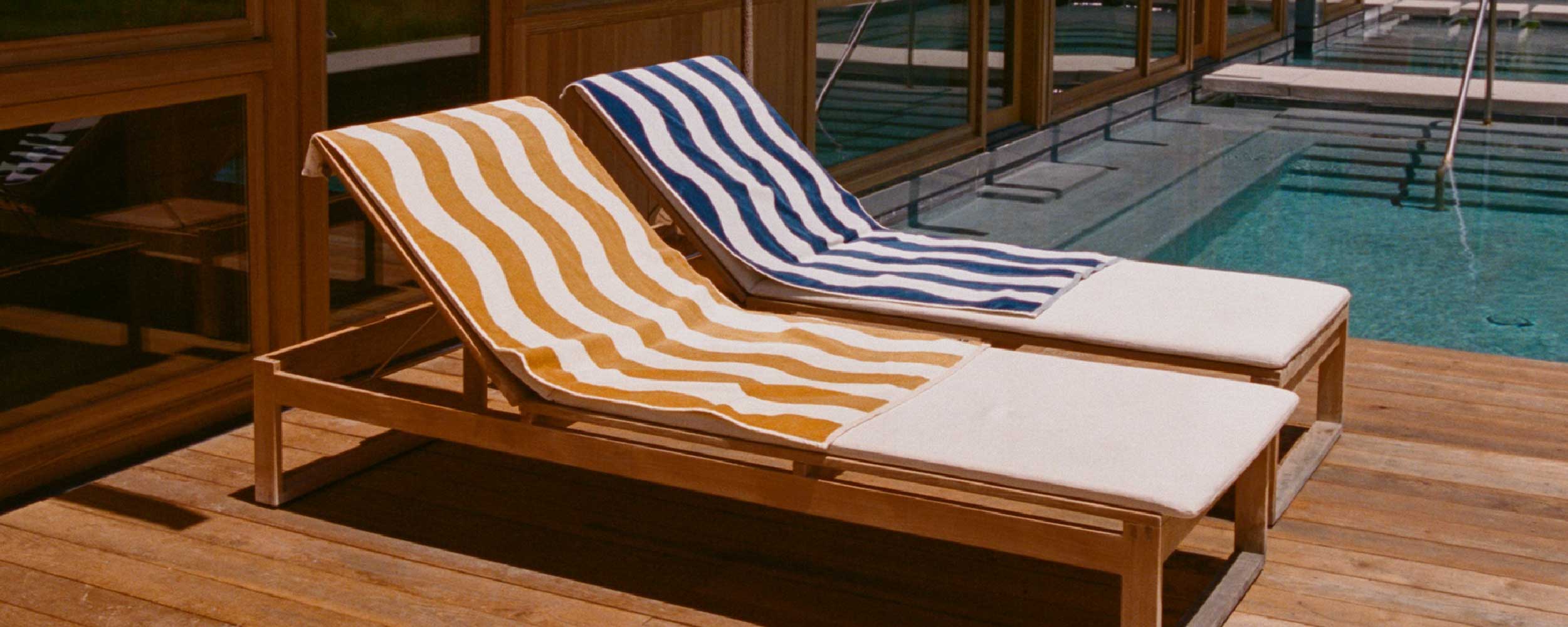 Two wooden lounge chairs with striped towels—one yellow, one blue—are placed side by side on a wooden deck next to a swimming pool, with building windows reflecting the scene.