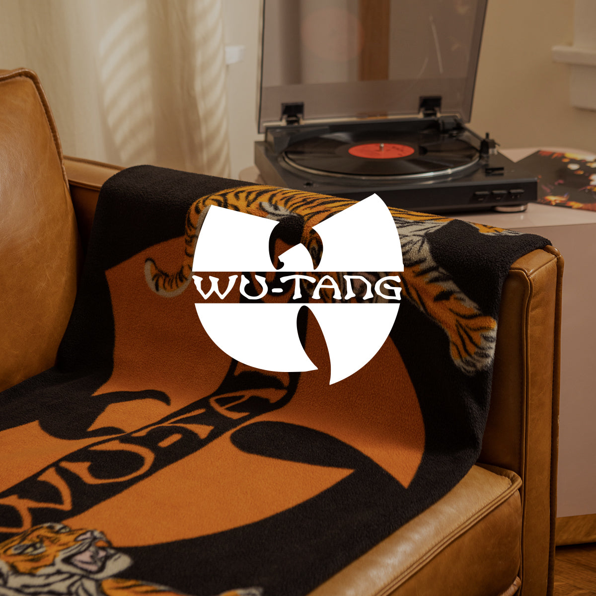 A brown leather chair draped with a Wu-Tang Clan blanket sits next to a small table holding a record player. The Wu-Tang Clan logo is prominently overlaid in the center of the image.