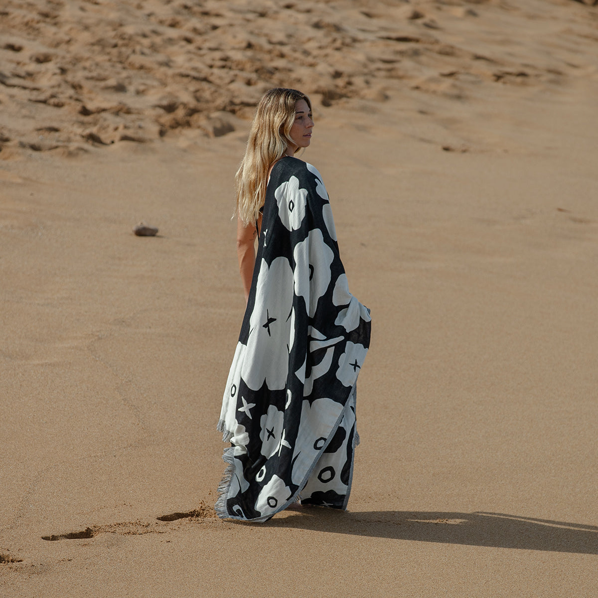 A woman with long blonde hair stands on a sandy beach, wrapped in a large black and white floral-patterned blanket, looking off into the distance.