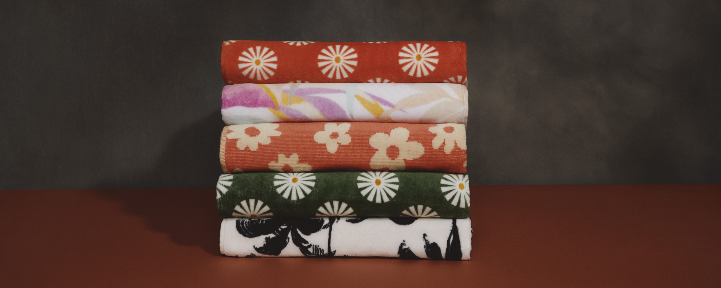 Five folded towels with various floral and abstract patterns in red, pink, orange, green, and black tones are stacked on a brown surface against a dark background.
