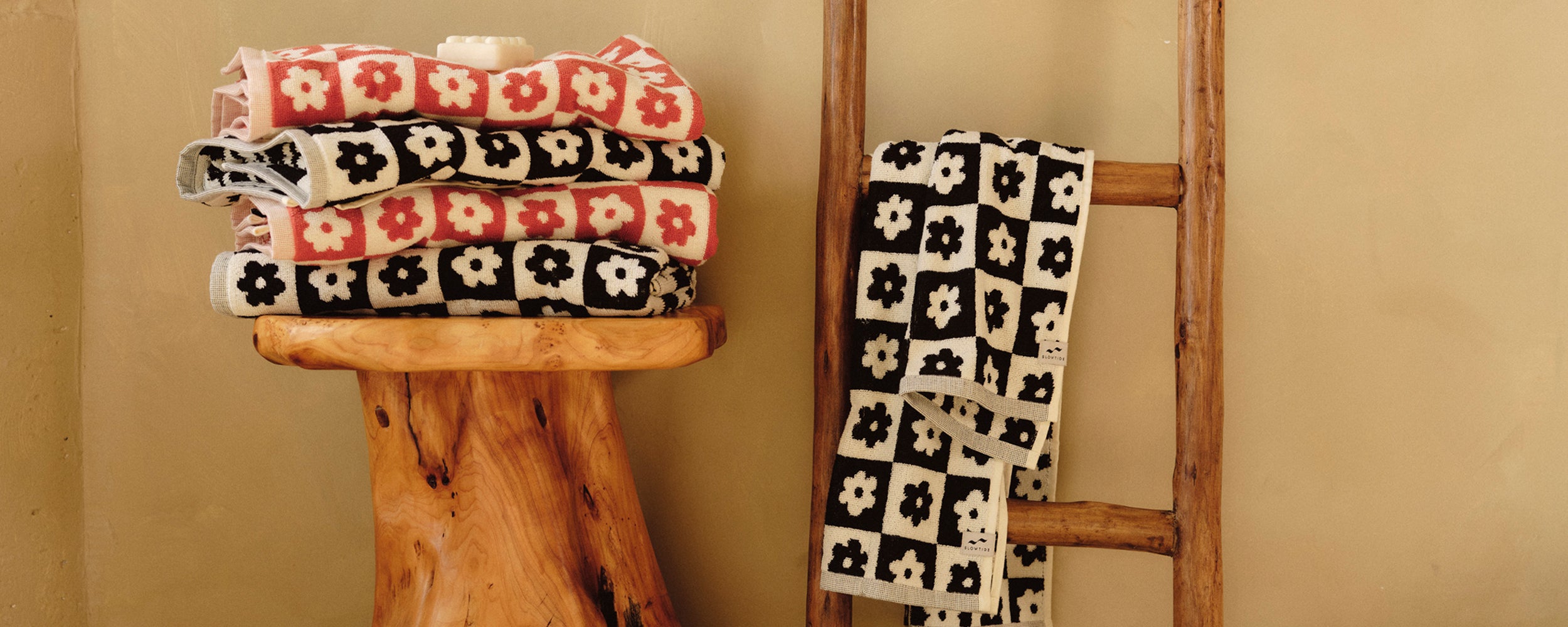 Two folded towels with red and white flower patterns sit on a wooden stool, while a black and white flower-patterned towel hangs on a rustic wooden ladder against a beige wall.