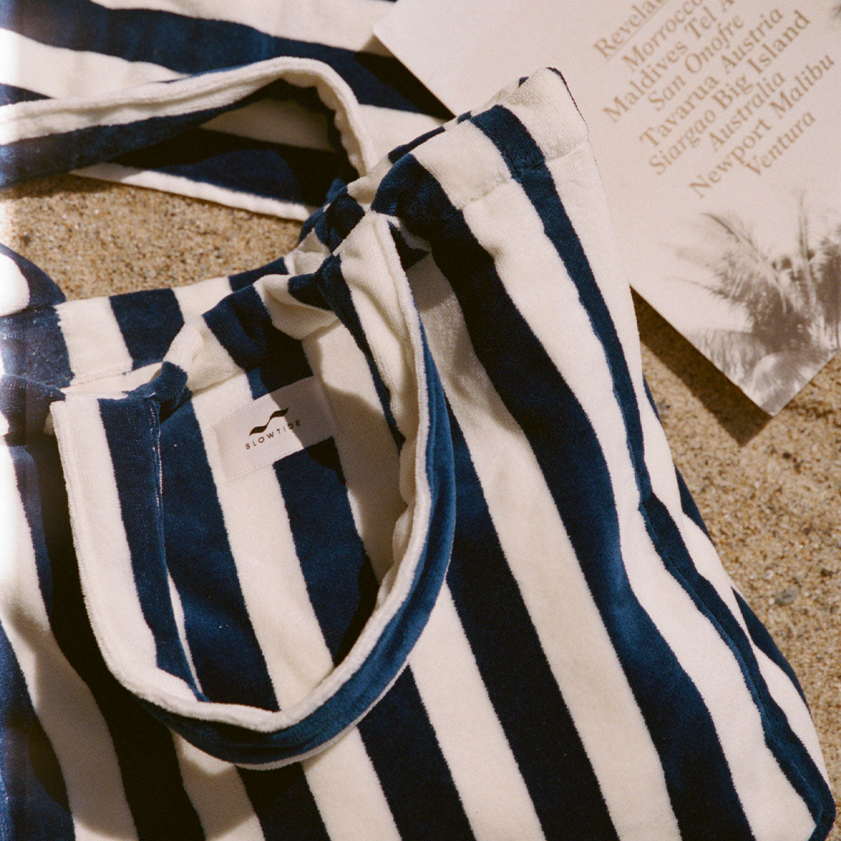 A blue and white striped tote bag rests on sand beside a piece of paper listing beach destinations, with a faint palm tree shadow visible on the paper.