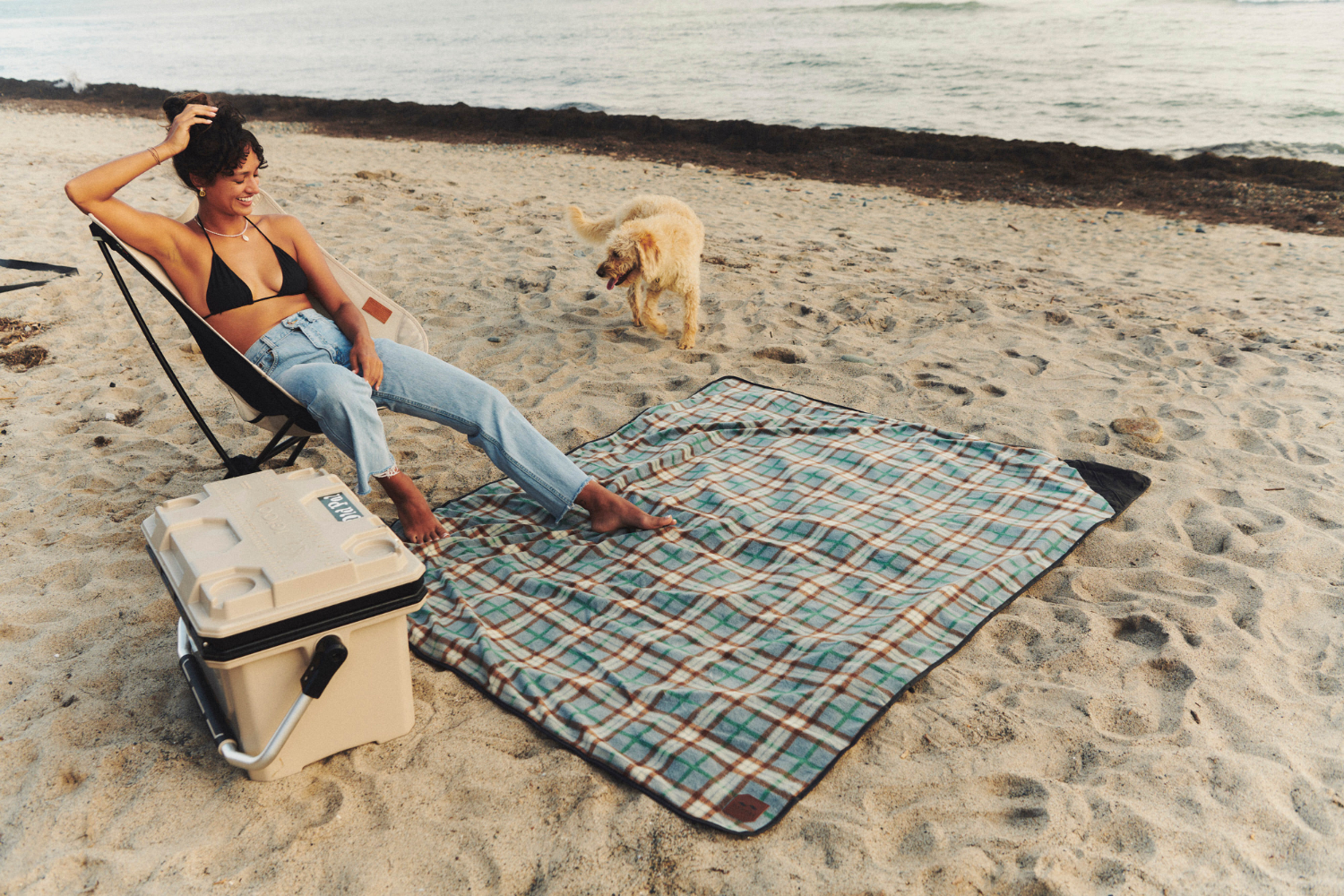 A woman in a black bikini top and jeans relaxes on a beach chair by a cooler and plaid blanket while a dog joyfully jumps nearby on the sandy shore by the ocean.