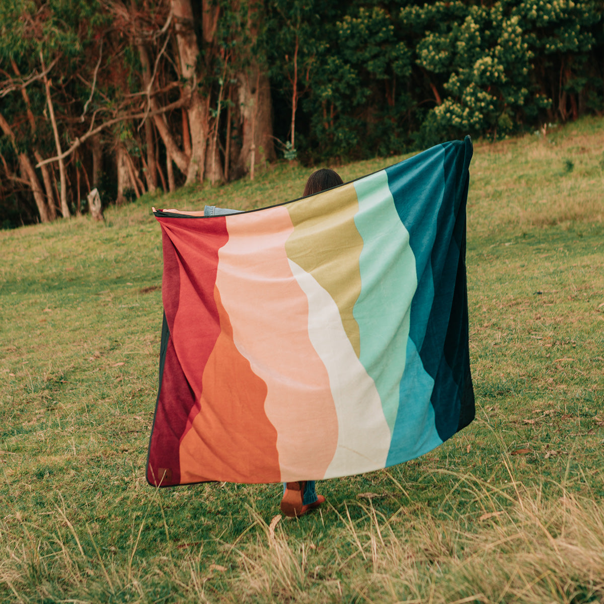 A person stands on grass holding a large, colorful blanket with wavy stripes in red, orange, yellow, green, blue, and navy, with trees and bushes in the background.