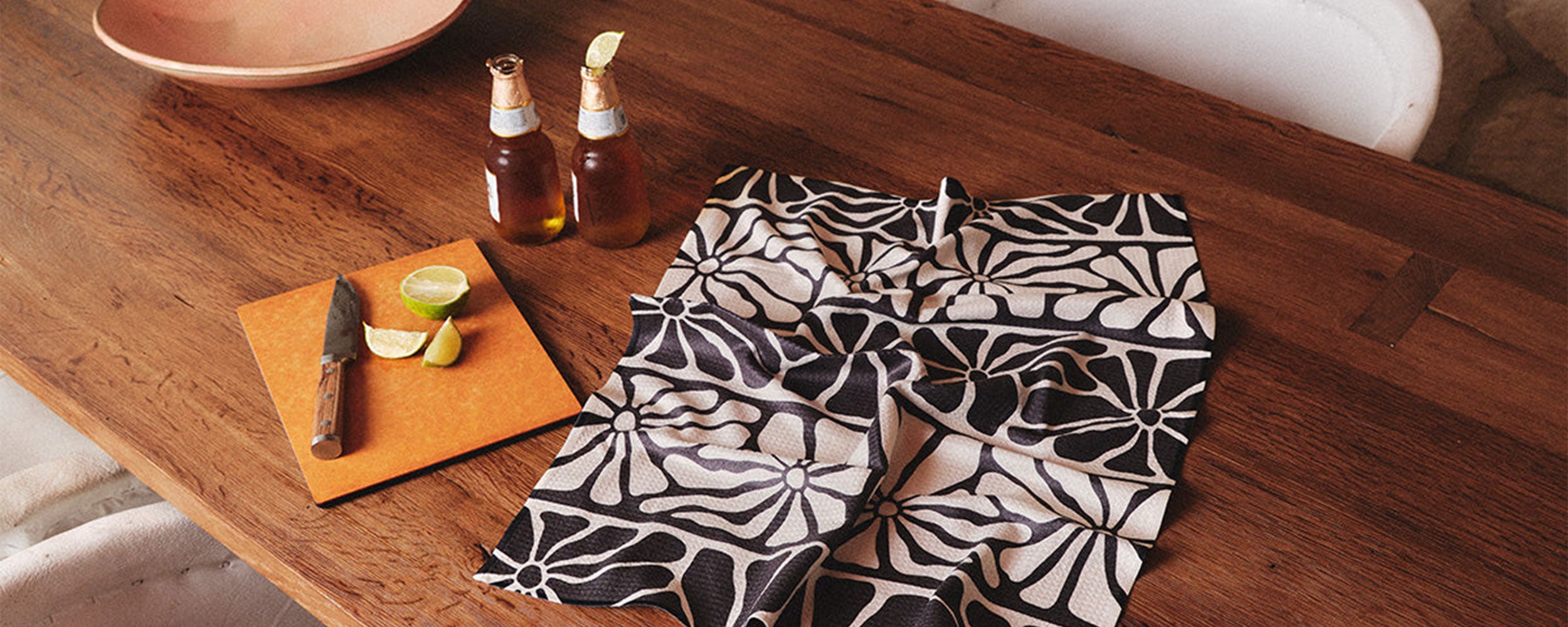 A wooden table with two beers, lime wedges on a cutting board with a knife, and a black and white patterned cloth spread out.
