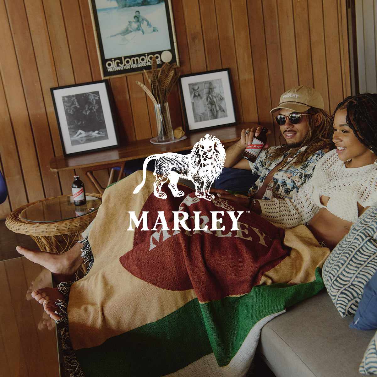 A smiling couple relaxes on a sofa under a Marley-branded blanket in a cozy wood-paneled room, with framed photos and a bottle on a nearby table.