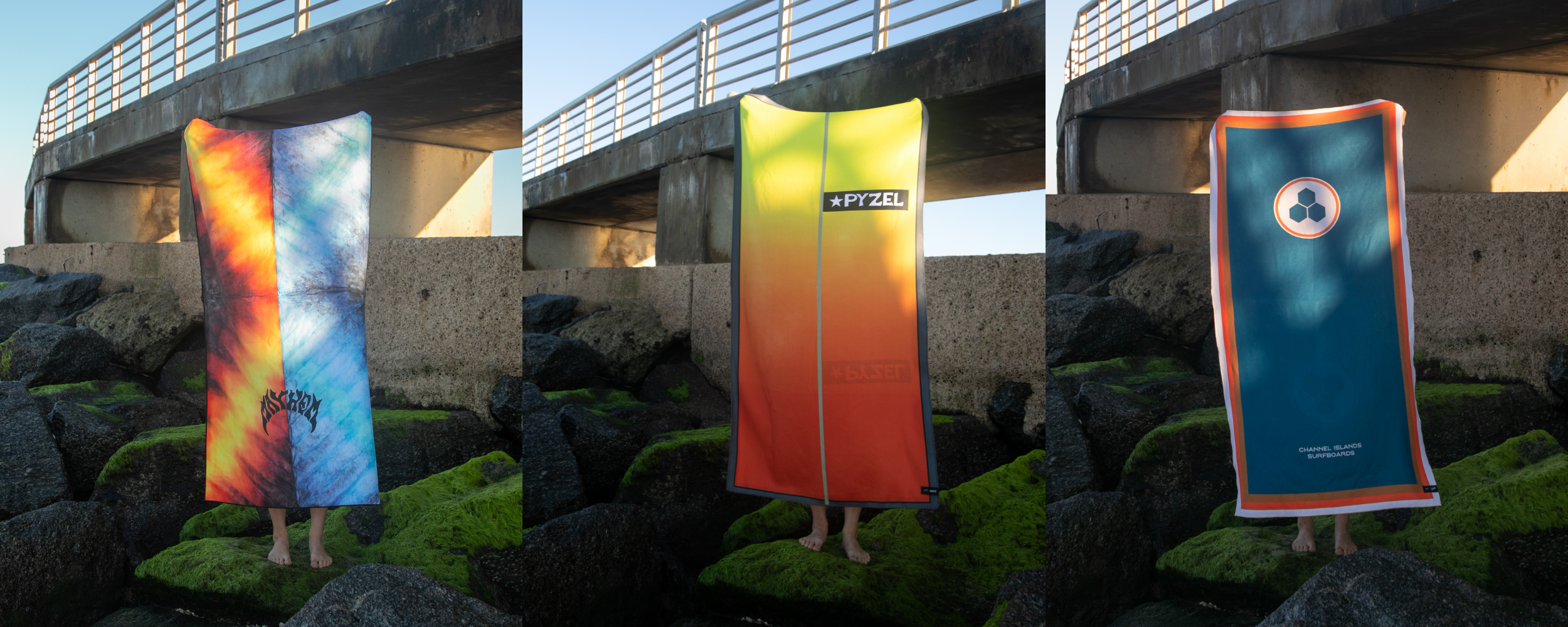 Three surfboards with colorful designs are held vertically on rocks under a pier, obscuring the people behind them, with a concrete wall and metal railing in the background.
