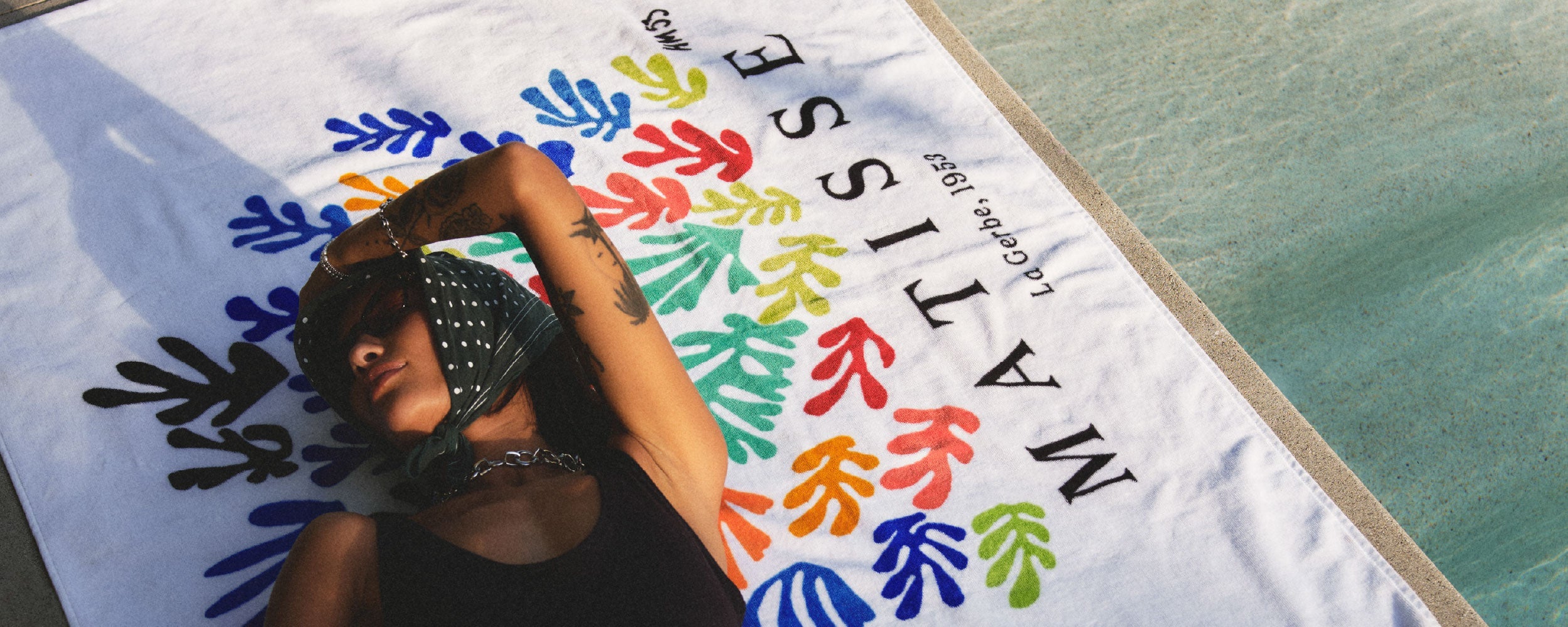 A person in sunglasses and a headscarf relaxes on a poolside towel featuring colorful leaf shapes and the text MATISSE La Gerbe, 1953.