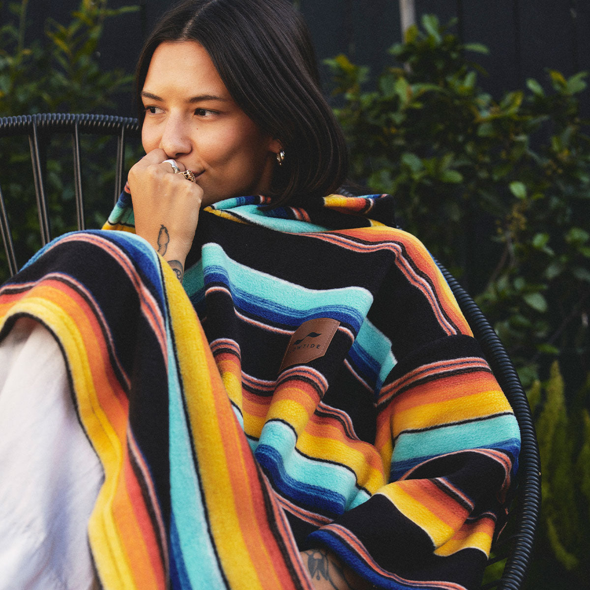 A person with dark hair sits outdoors on a chair, wrapped in a colorful striped blanket. They are smiling softly, with one hand near their face, and greenery is visible in the background.