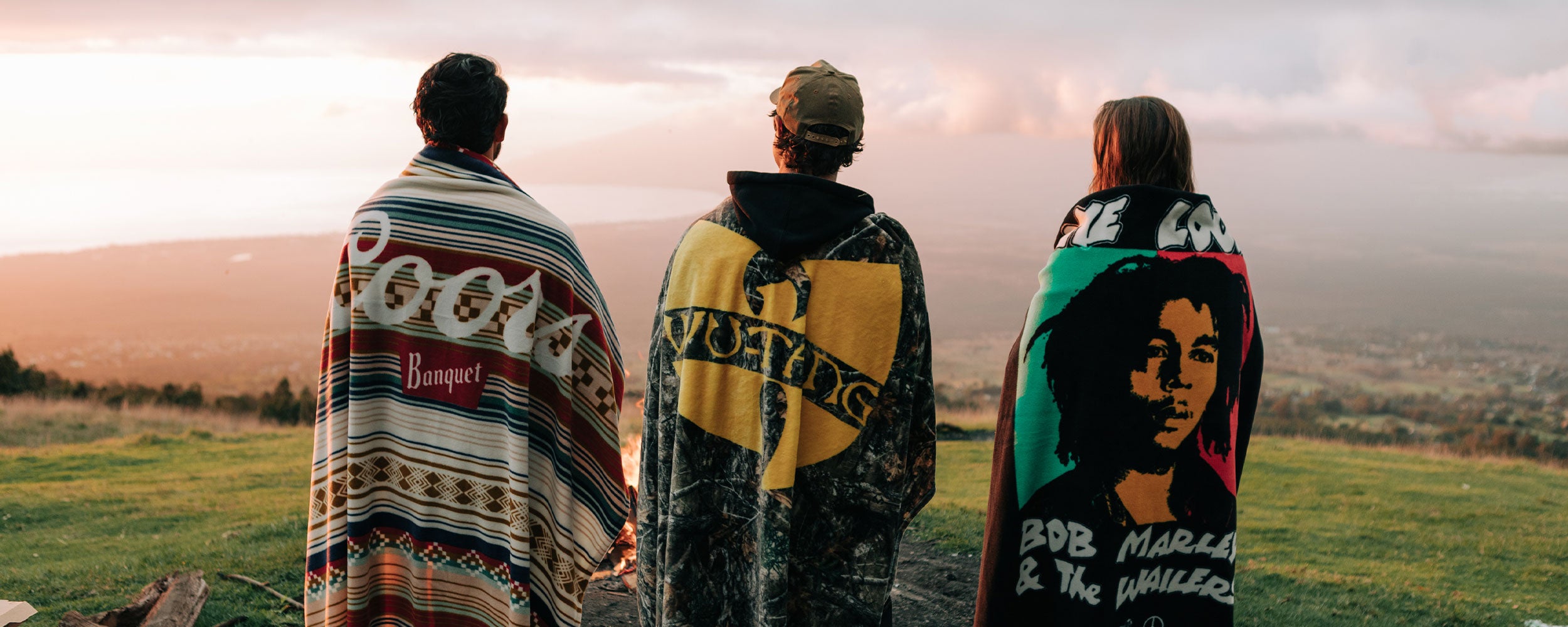 Three people stand outdoors, wrapped in blankets, facing a scenic landscape at sunset. The blankets feature colorful designs, including one with Bob Marley’s portrait.