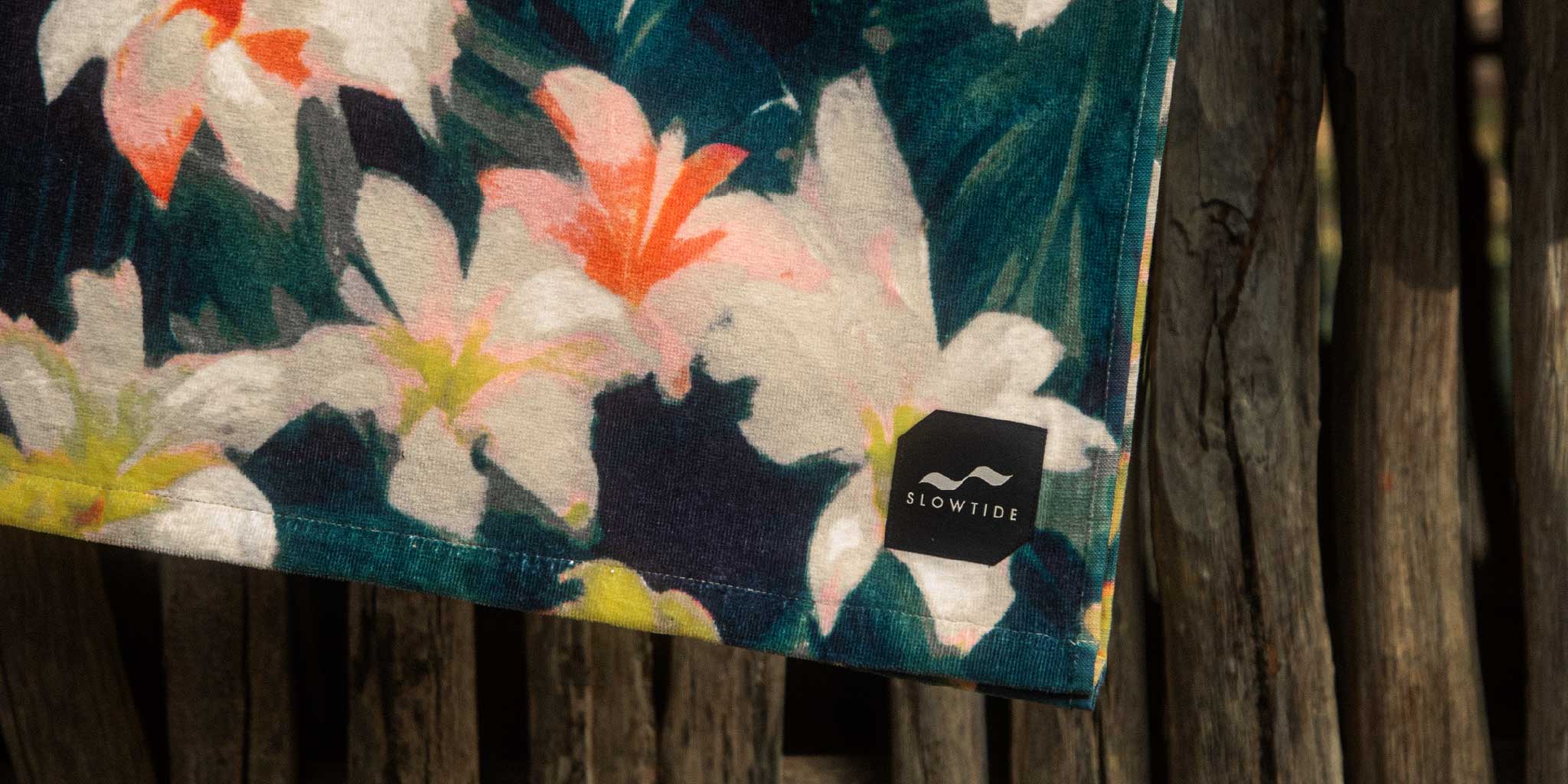A close-up of a colorful floral-patterned fabric with white and orange flowers, featuring a small black SLOWTIDE tag, hanging in front of wooden slats.