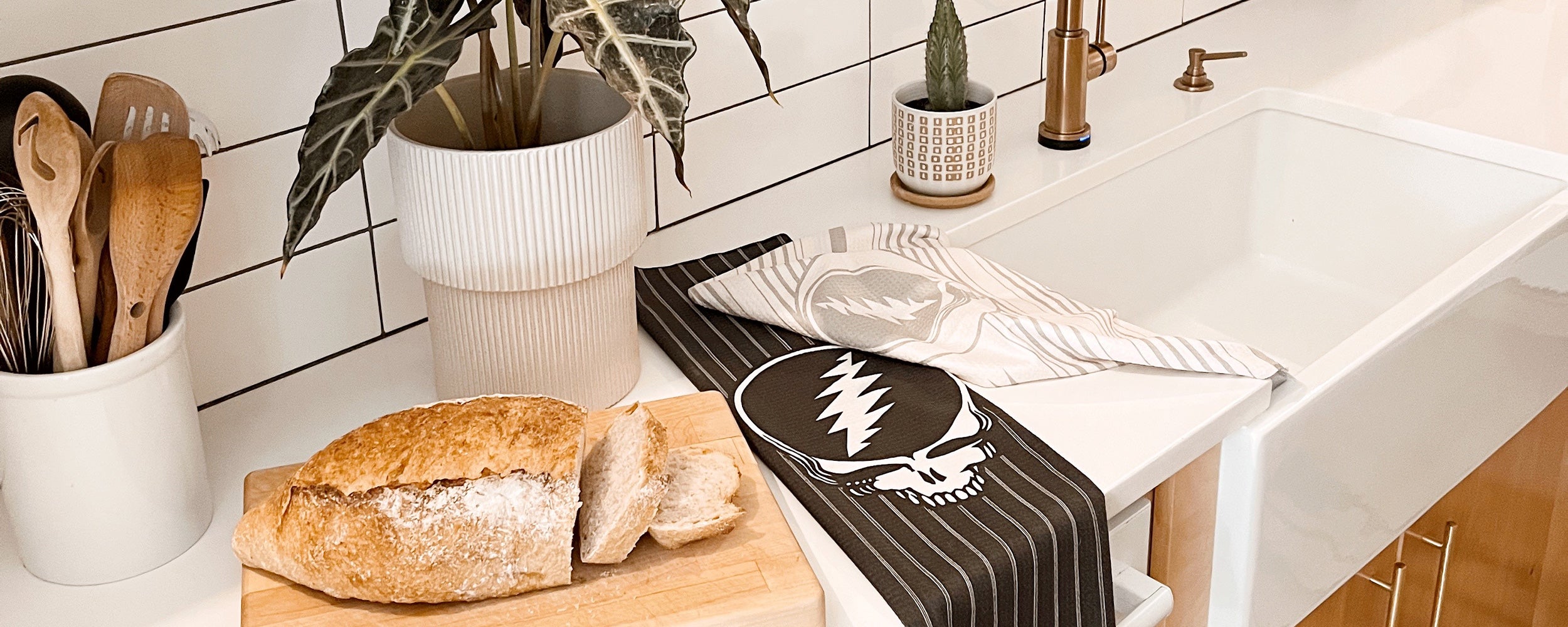 A rustic loaf of bread, partially sliced on a cutting board, sits by a kitchen sink with potted plants, wooden utensils in a holder, and two dish towels draped on the counter.