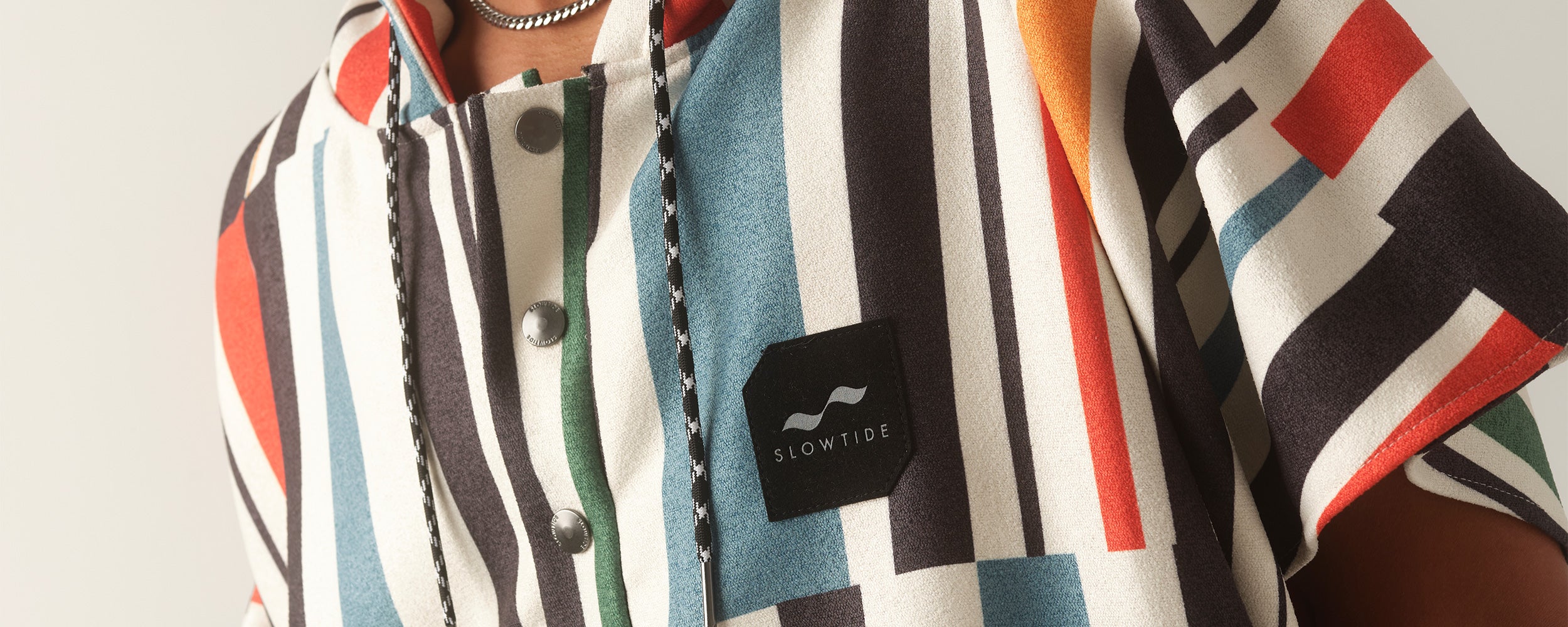 A person wearing a colorful, geometric-patterned shirt with a Slowtide patch on the chest and black-and-white braided drawstrings.
