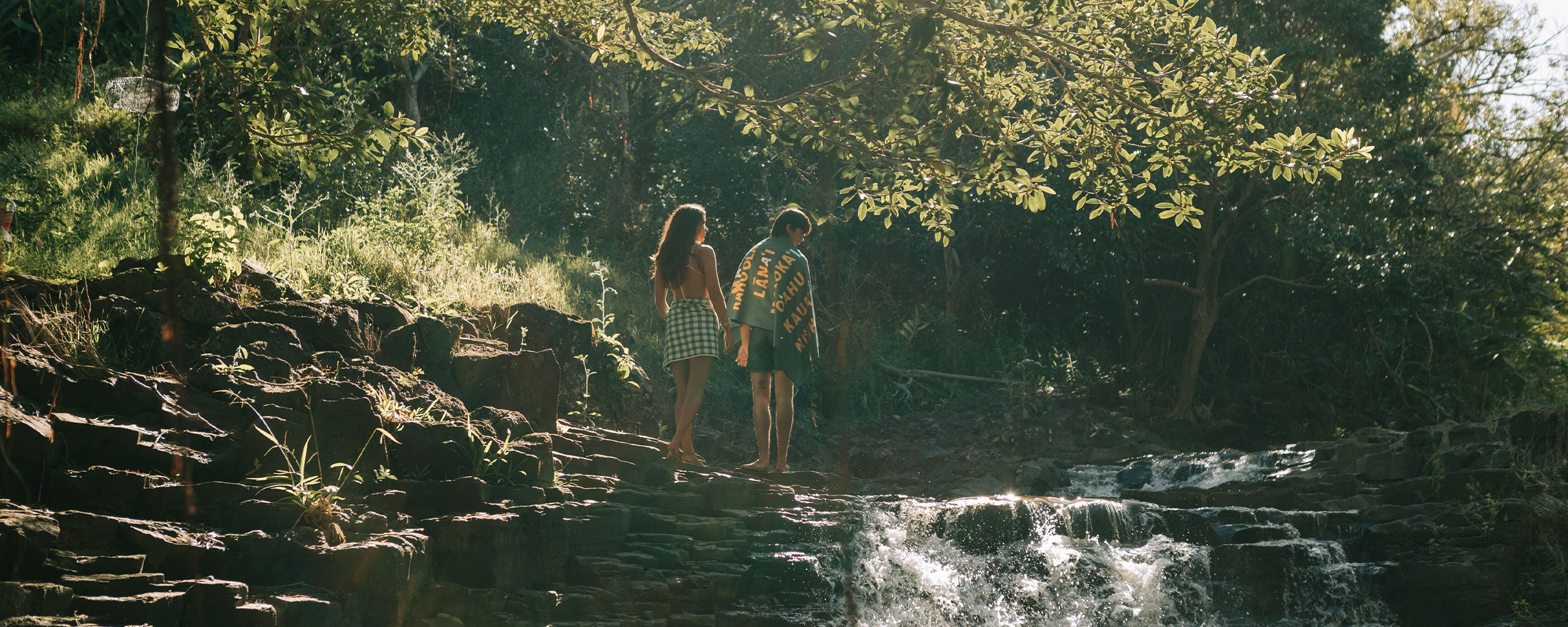 Two people holding hands walk along rocky terrain beside a gentle waterfall, surrounded by lush greenery and sunlight filtering through the trees.