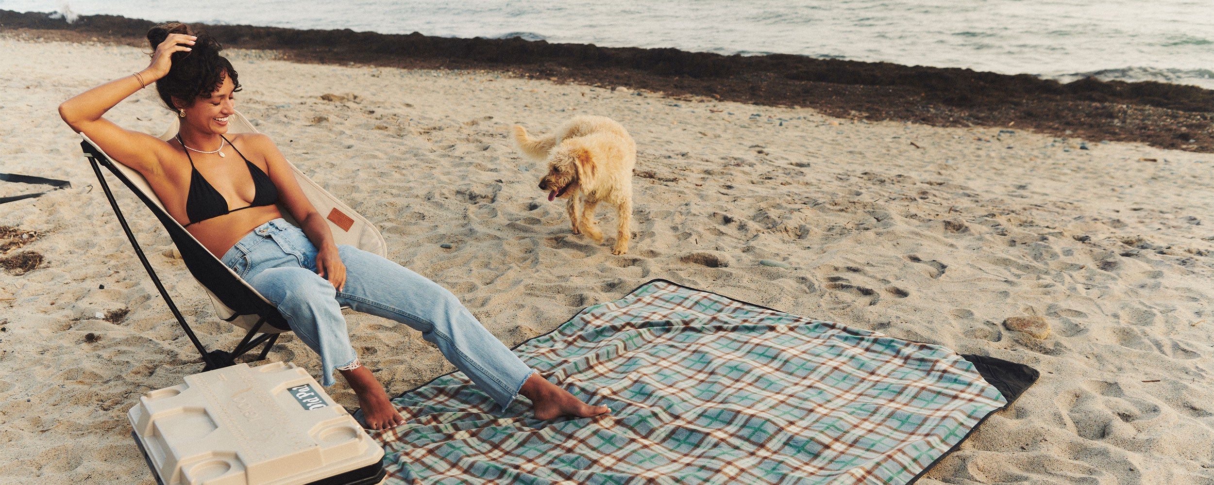 A woman in a black bikini top and jeans relaxes on a beach chair by a plaid blanket, smiling at a playful dog running on the sandy shore near the ocean.