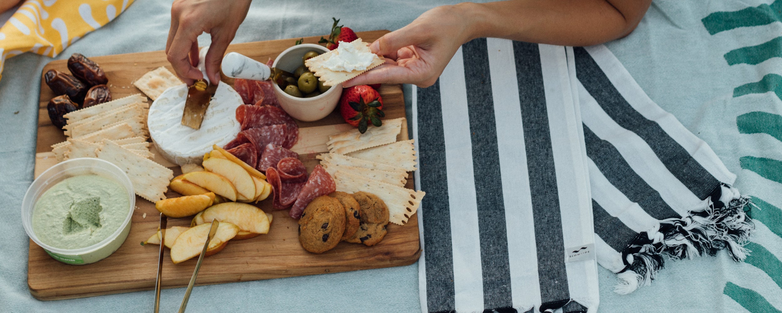 A hand spreads cheese on a cracker over a wooden board with salami, crackers, cookies, apple slices, strawberries, olives, dates, and dip, next to a striped towel on a picnic blanket.