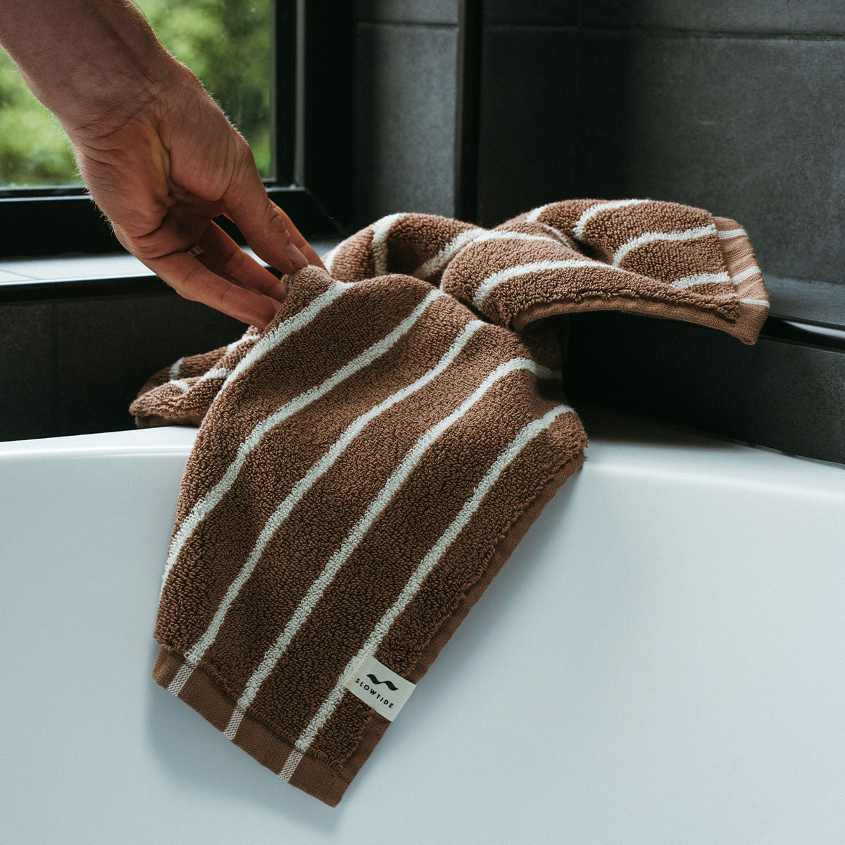 A hand reaches for a brown towel with white stripes draped over the edge of a white bathtub in a modern bathroom.