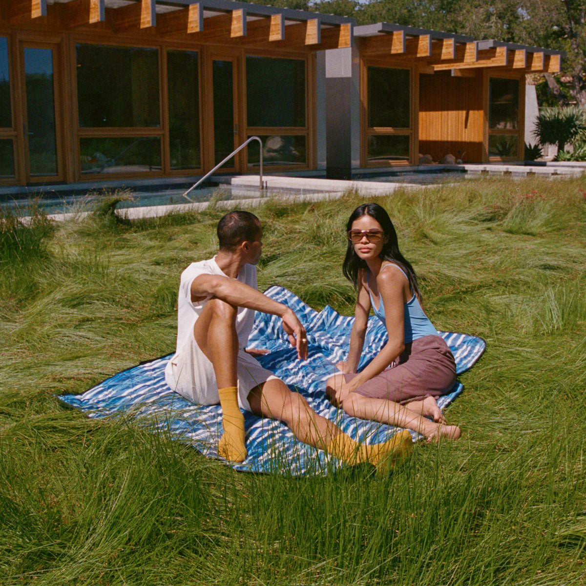Two people sit on a striped blanket in tall grass outside a modern wooden house, enjoying a sunny day. One person looks at the other, who gazes toward the camera, both dressed casually.