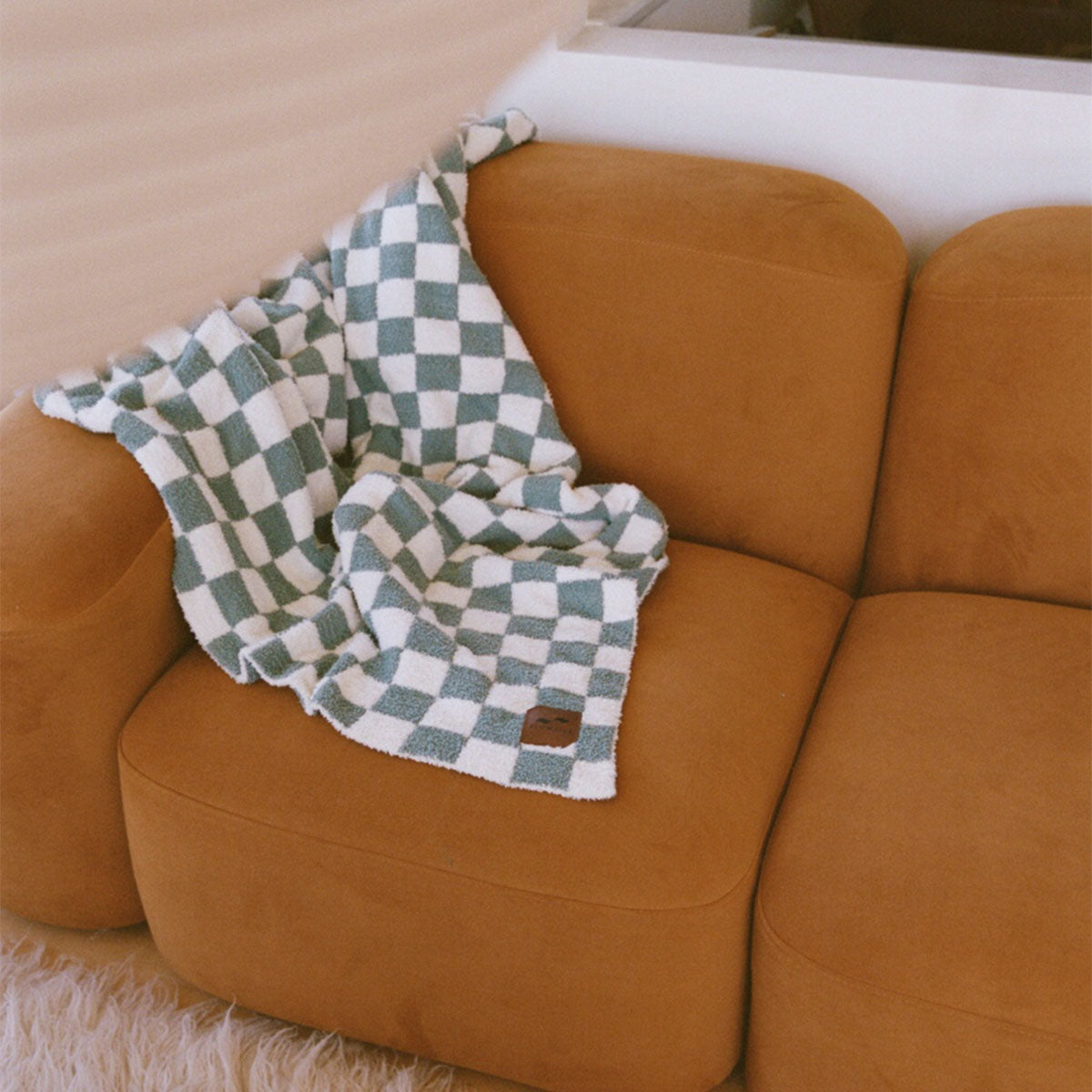 A soft, tan couch with a green and white checkered blanket draped over the armrest, next to a fluffy white rug and white walls in a cozy living room.