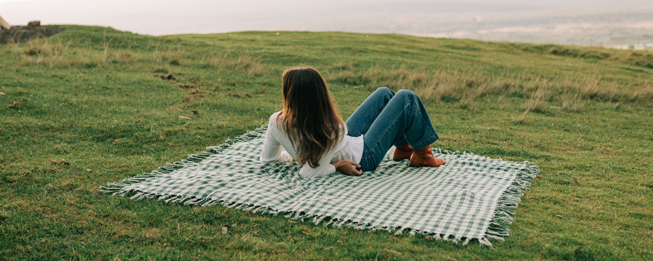 A woman with long hair relaxes on a checkered blanket laid out on a grassy hill, gazing into the distance. She wears a white top, blue jeans, and brown shoes. The sky is hazy in the background.