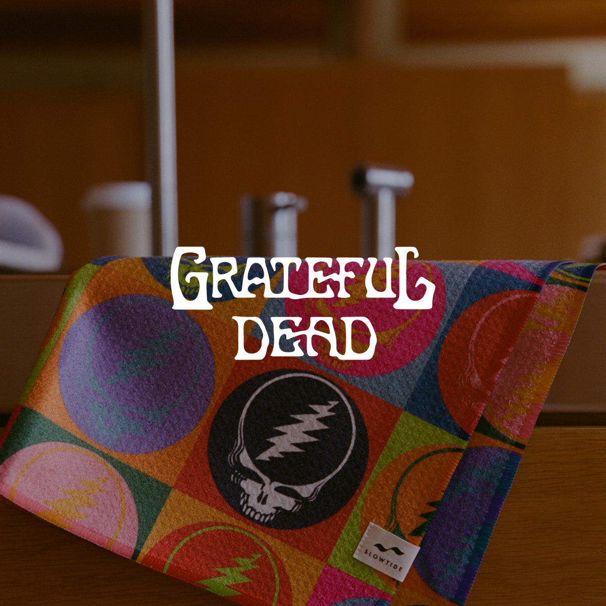 A colorful towel with Grateful Dead logos and patterns draped over a counter. The words Grateful Dead appear in bold, white, retro-style font in the center.