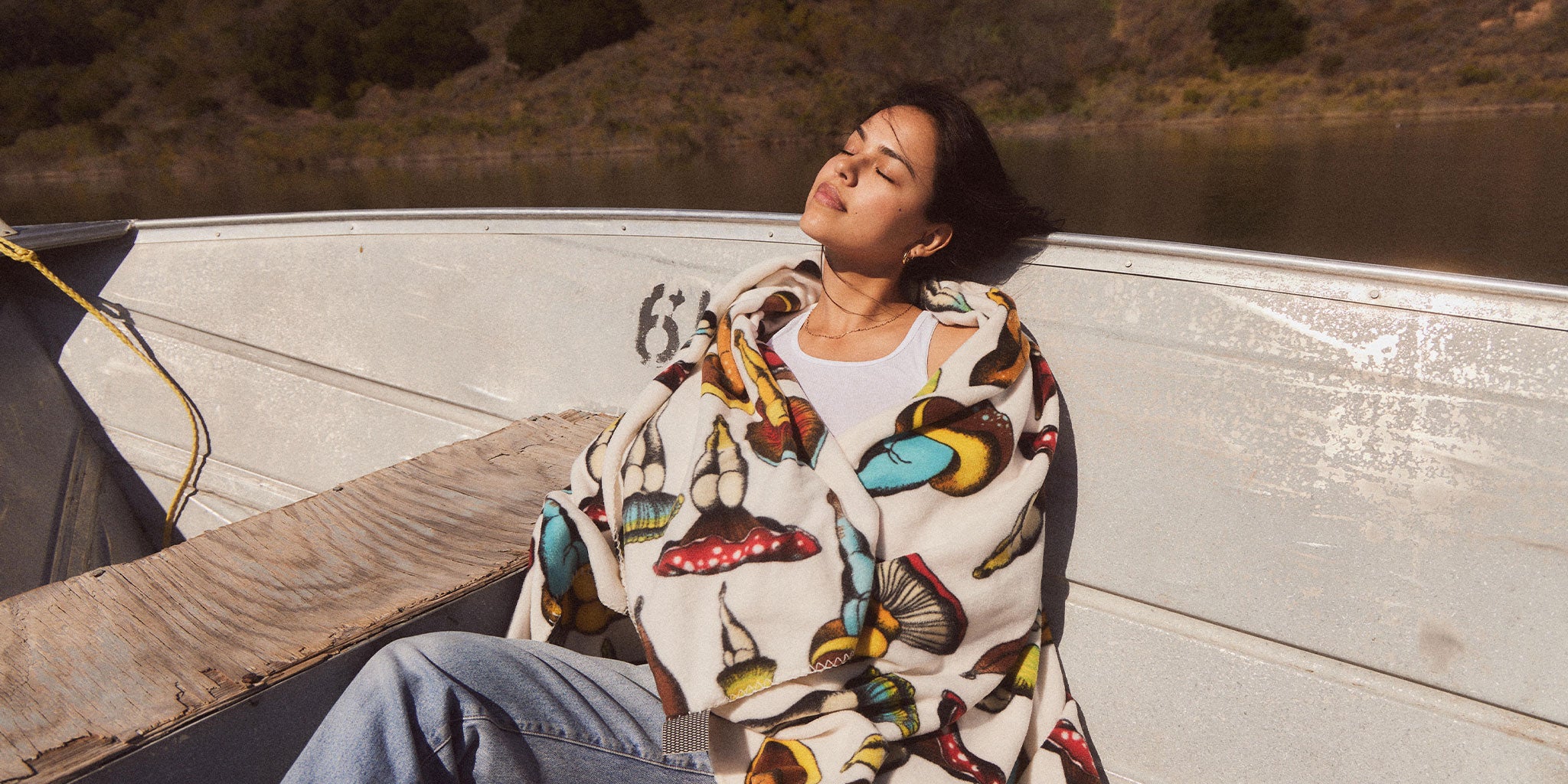 A woman sits on a boat, eyes closed, basking in the sun. She is wrapped in a blanket with colorful mushroom patterns, with calm water and greenery in the background.